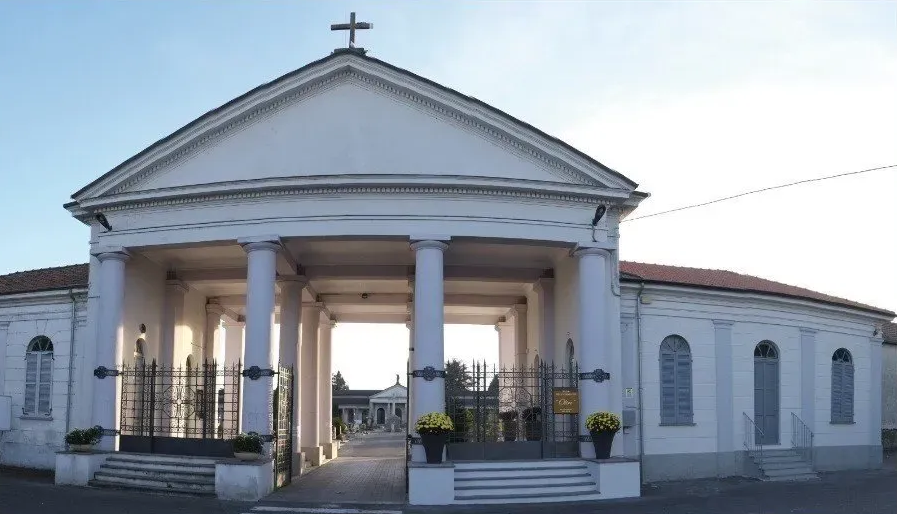 Ingresso bianco del cimitero in stile neoclassico con colonne, croce e cancelli in ferro battuto.