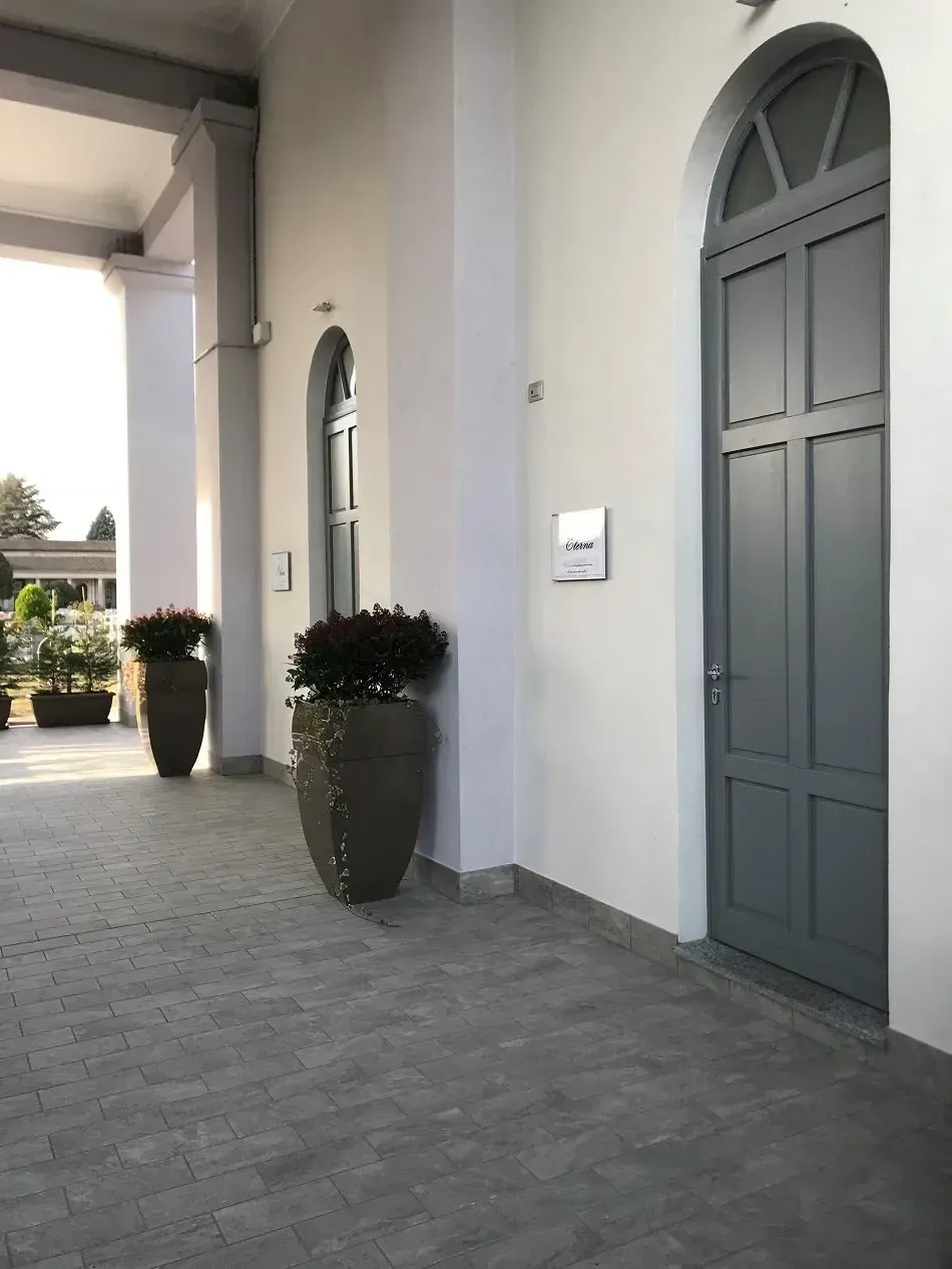 Facciata grigia dell'edificio con porta e finestra ad arco, fioriere con fogliame verde e un passaggio coperto.