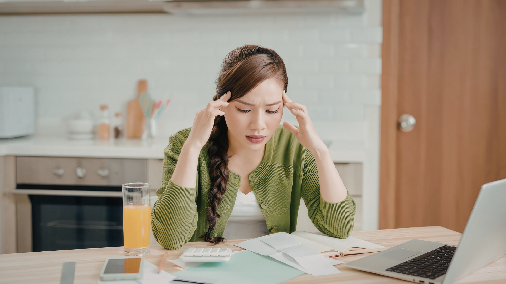 Woman sitting at a kitchen table, holding her temples while looking stressed over paperwork and a laptop