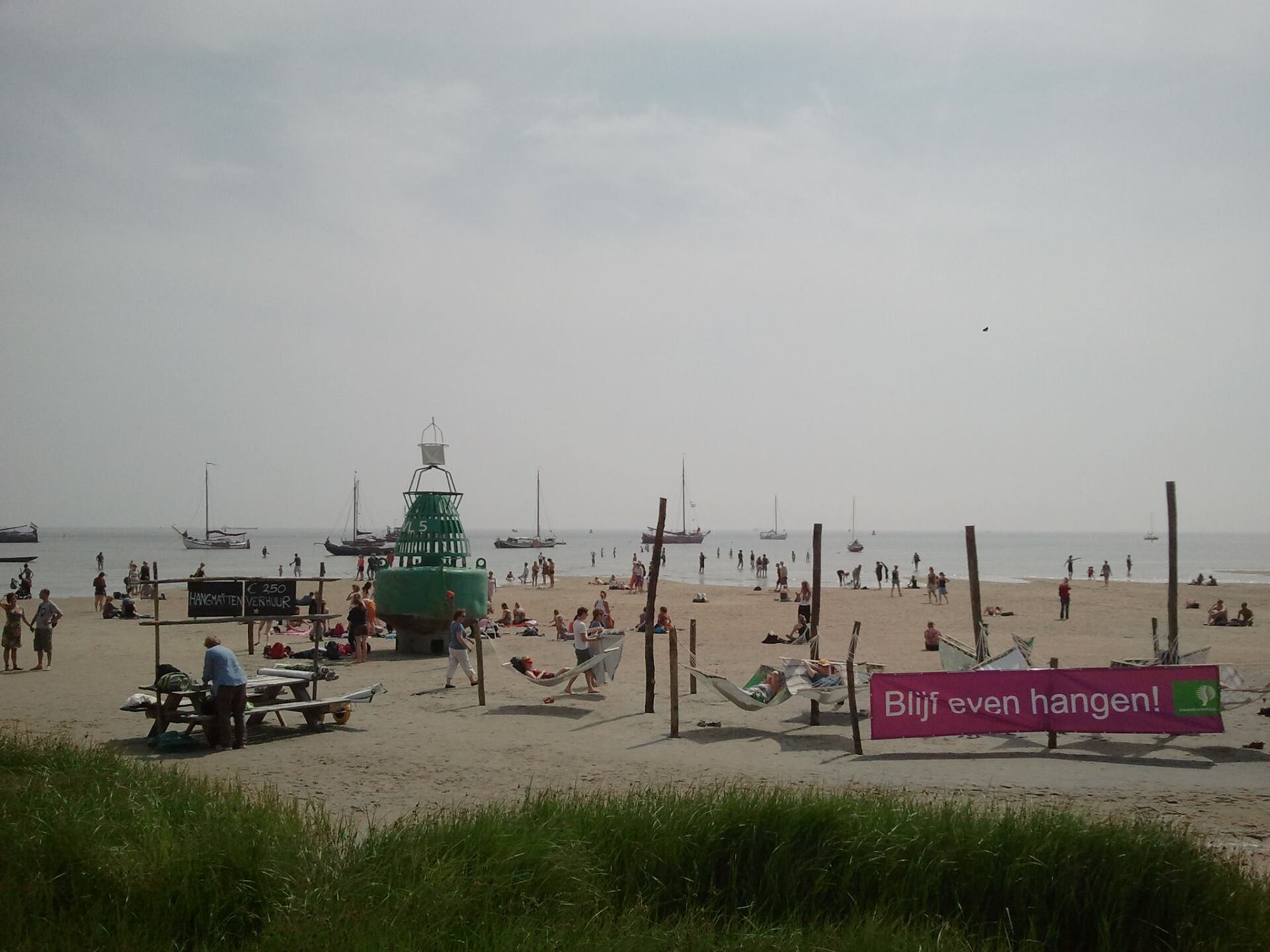 Strandszene mit Menschen, Segelbooten, grüner Boje, Hängematten und Schild.