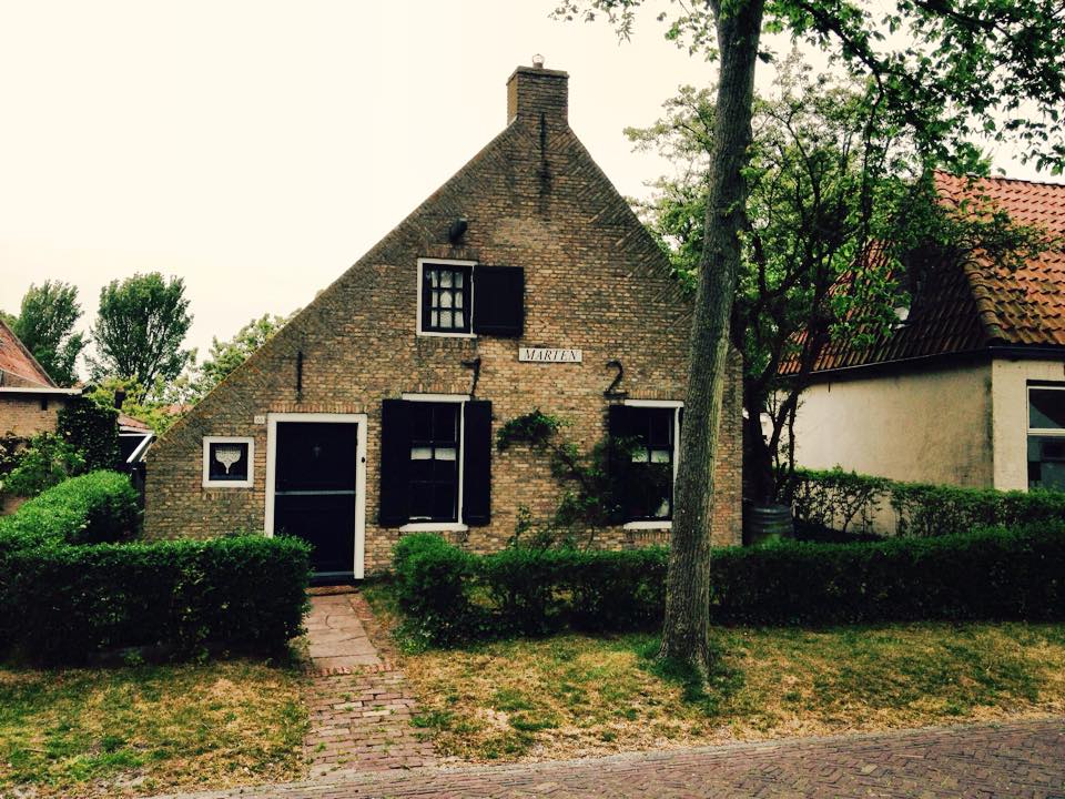 Steinhaus mit dunkler Tür und Fensterläden, einem kleinen quadratischen Fenster und geschnittenen Hecken davor.