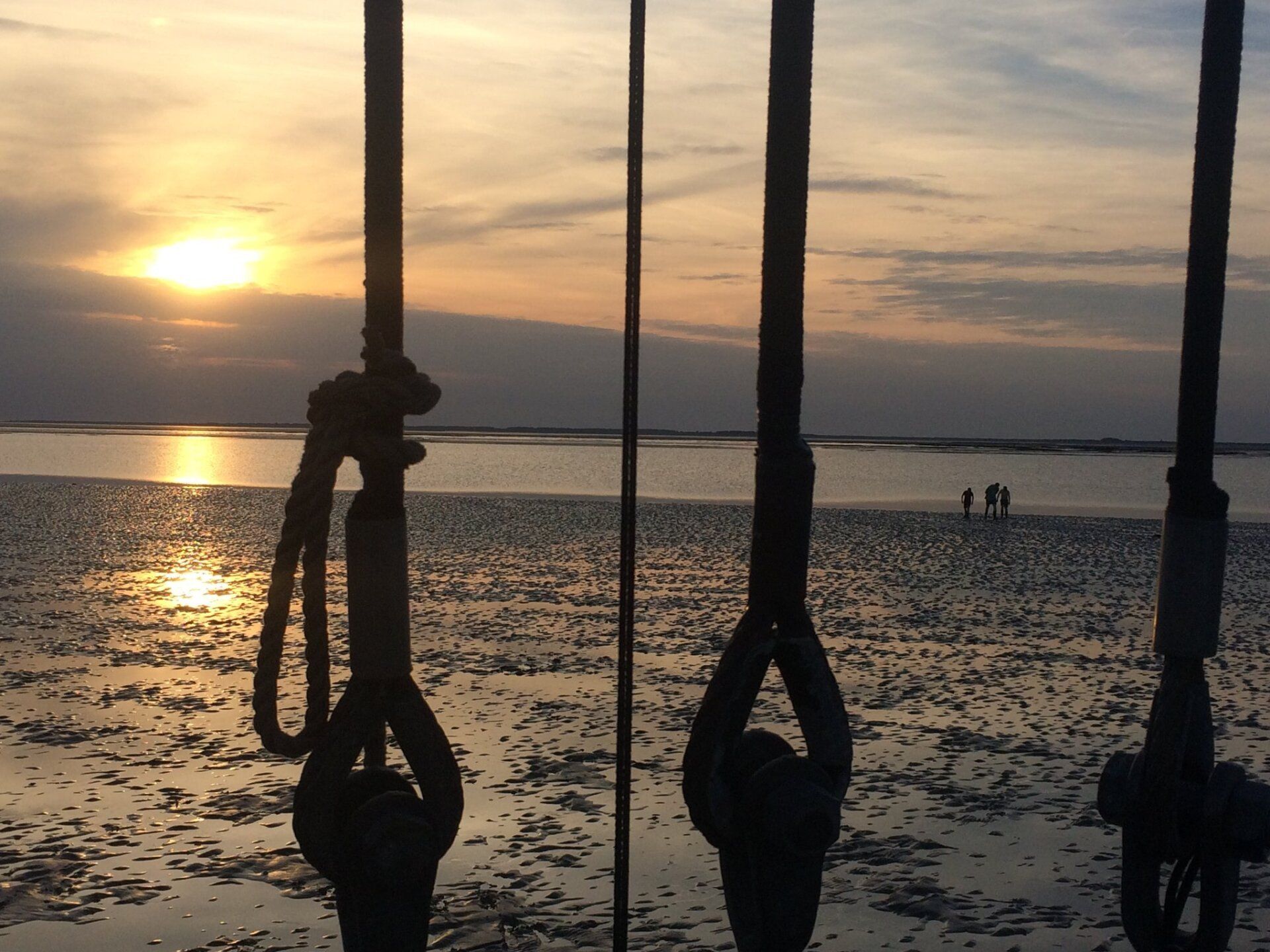 Sonnenuntergang über einem Strand, durch die Takelage betrachtet. Silhouetten von Figuren gehen am Ufer entlang.