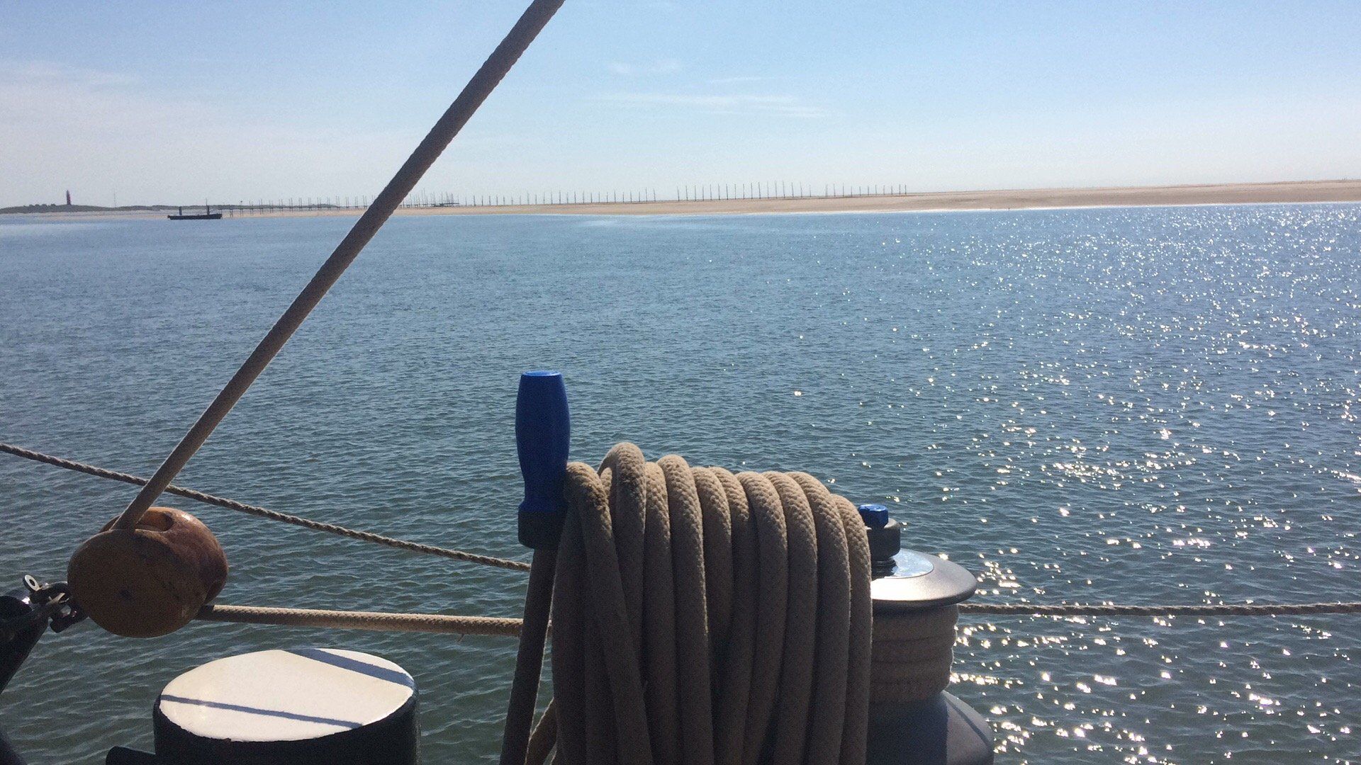 Bootsdeck mit aufgerolltem Seil, mit Blick auf glitzerndes Wasser, Küste und blauen Himmel.