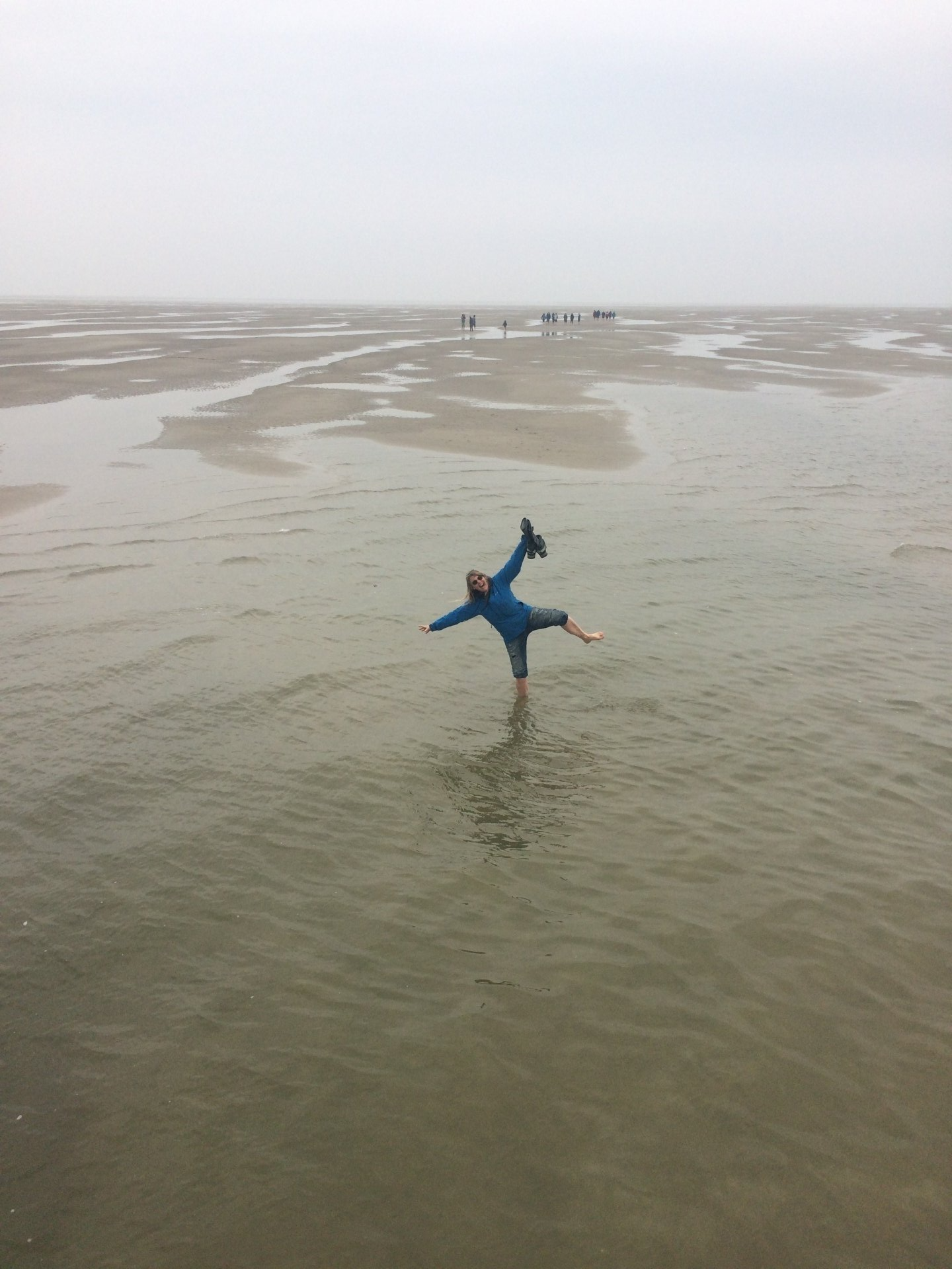 Person in Blau steht im seichten Wasser und balanciert mit einem Bein auf einem flachen, nassen Strand. Bewölkter Himmel.