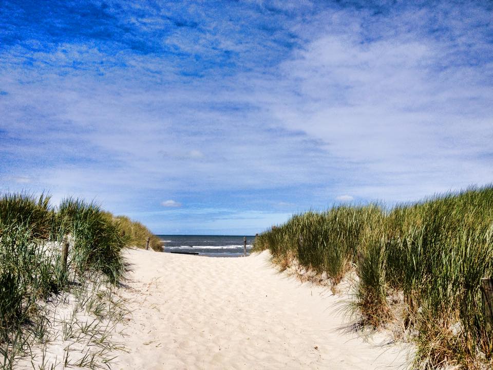Ein Sandweg führt zwischen grasbewachsenen Dünen unter einem blauen Himmel mit dünnen Wolken zum Meer.