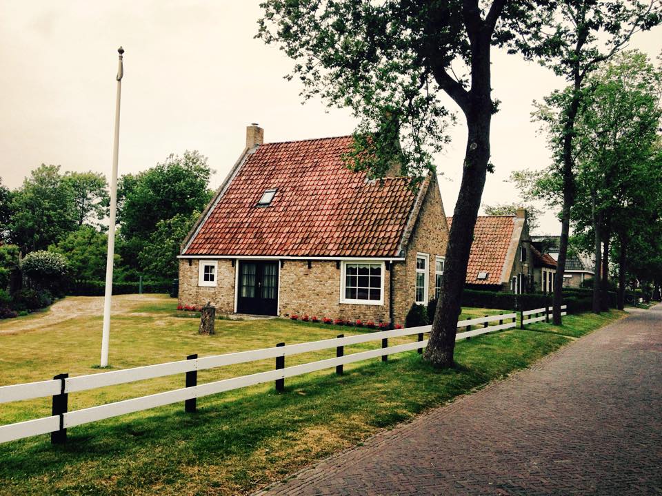Haus mit rotem Ziegeldach, weißem Lattenzaun und Fahnenmast auf einer Rasenfläche neben einer Straße, bewölkter Himmel.