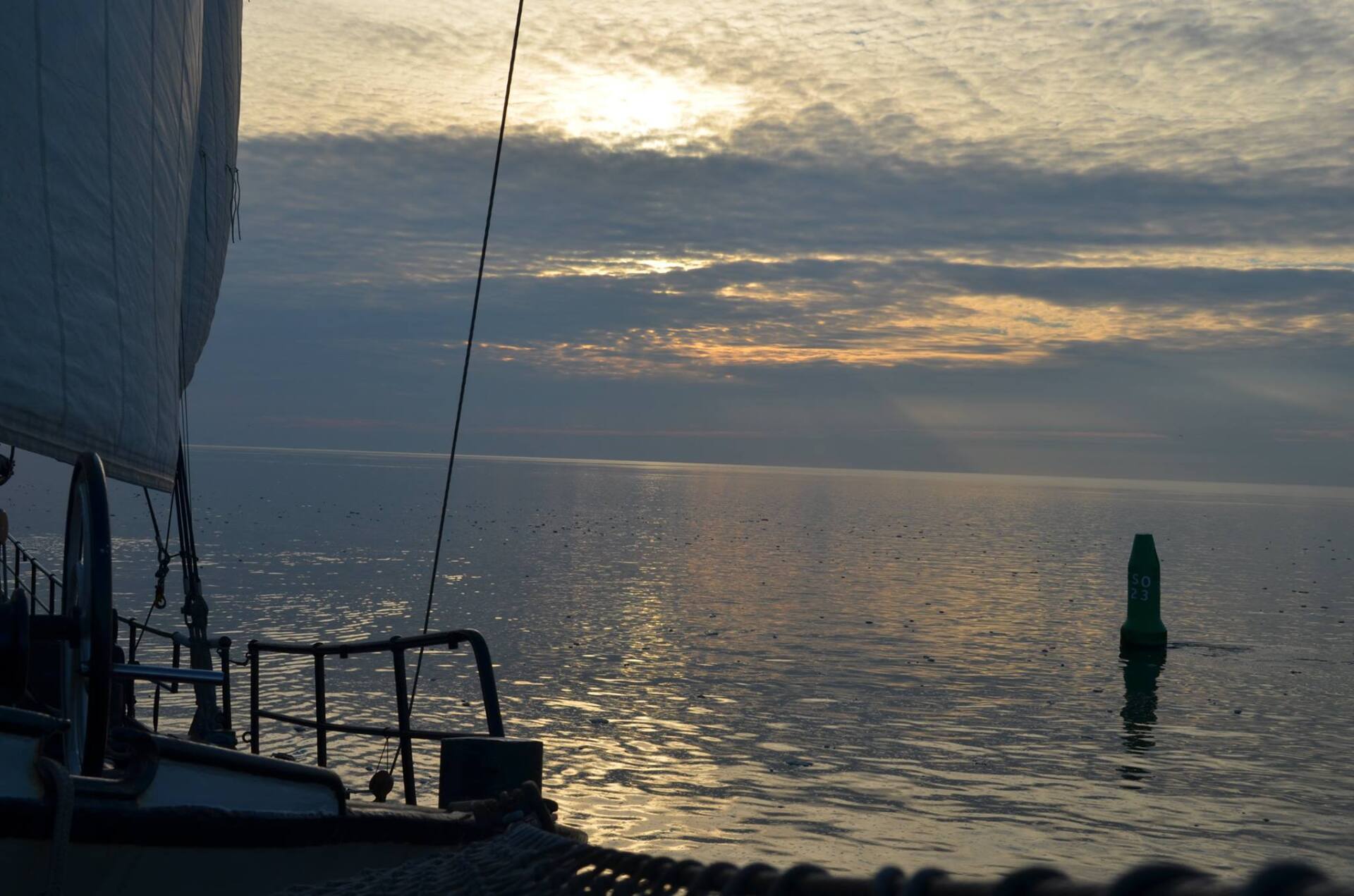 Segelboot segelt bei Sonnenaufgang auf ruhigem Wasser, in der Ferne ist eine grüne Boje zu sehen.