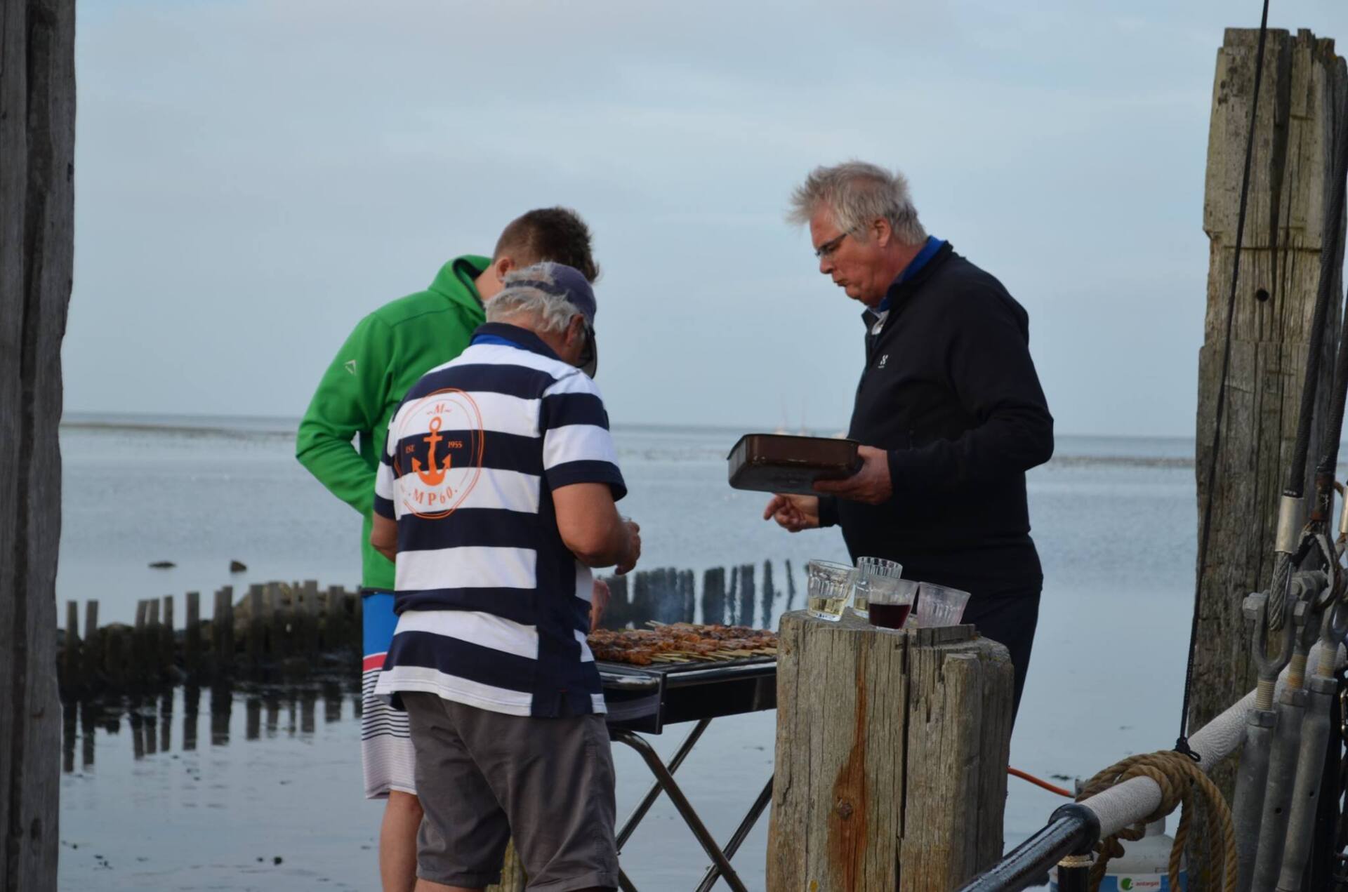 Drei Männer grillen im Freien in der Nähe von Wasser; bewölkter Himmel; ein Mann hält Essen.