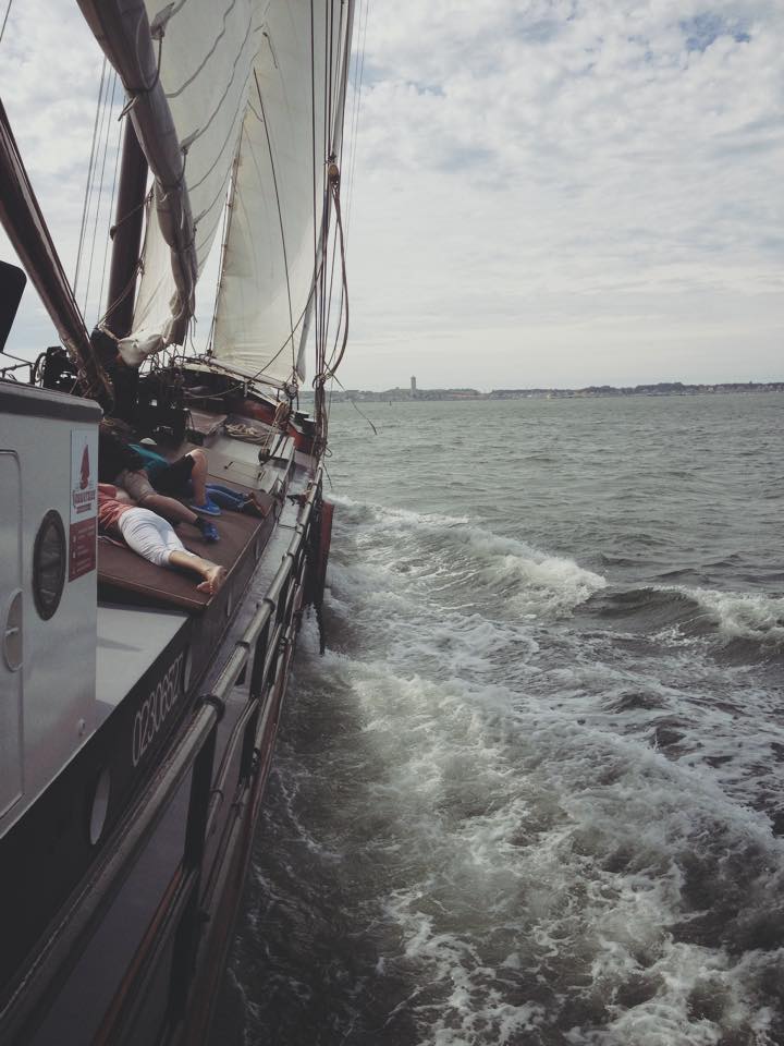 Segelboot auf dem Wasser, Wellen schlagen an die Seite. Menschen an Deck, bewölkter Himmel, entfernte Stadtlandschaft.