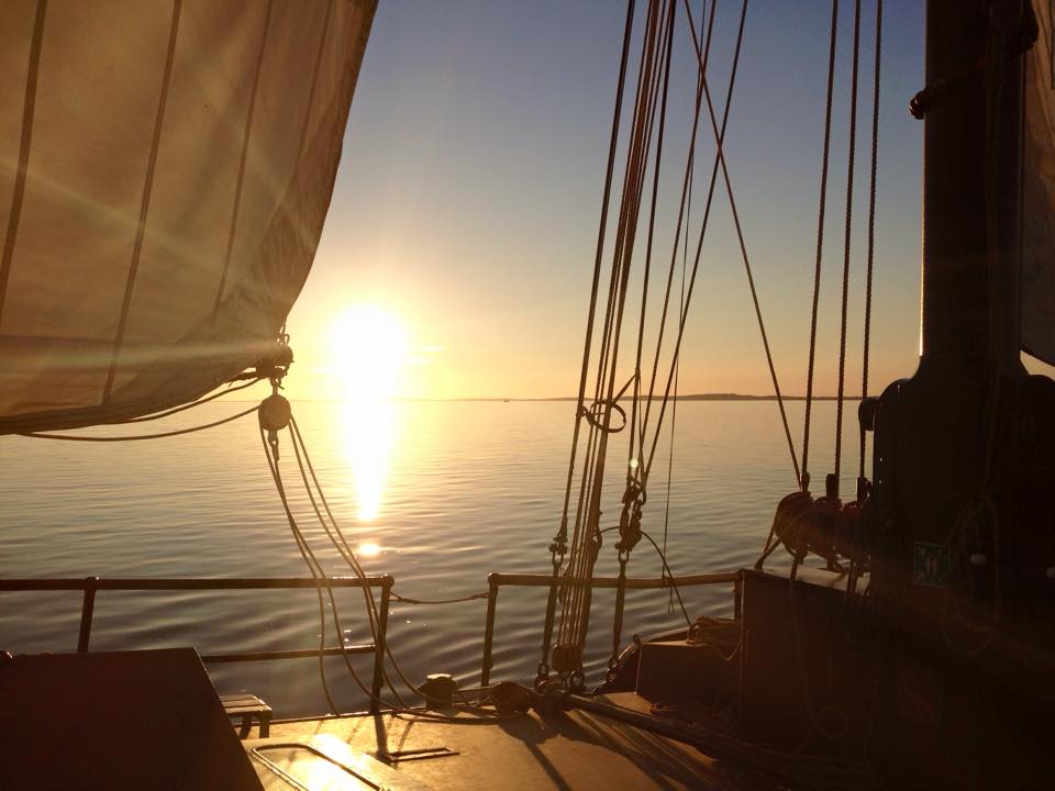 Sonnenuntergang über ruhigem Wasser, vom Deck eines Segelboots aus gesehen, Sonne reflektiert, Seile und Segel sichtbar.