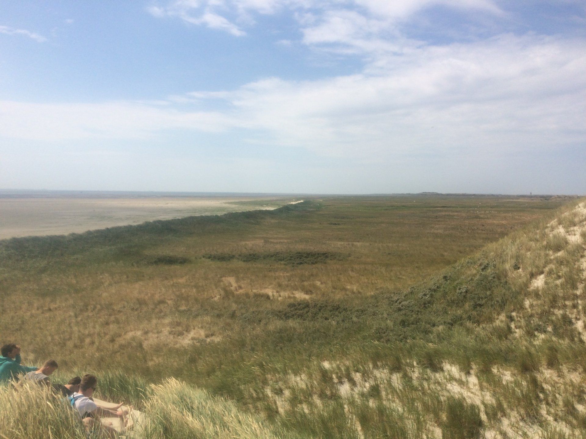 Grasbewachsene Dünen mit Blick auf eine weite, flache Fläche mit Wasser in der Ferne. Mehrere Personen sitzen am Hang.
