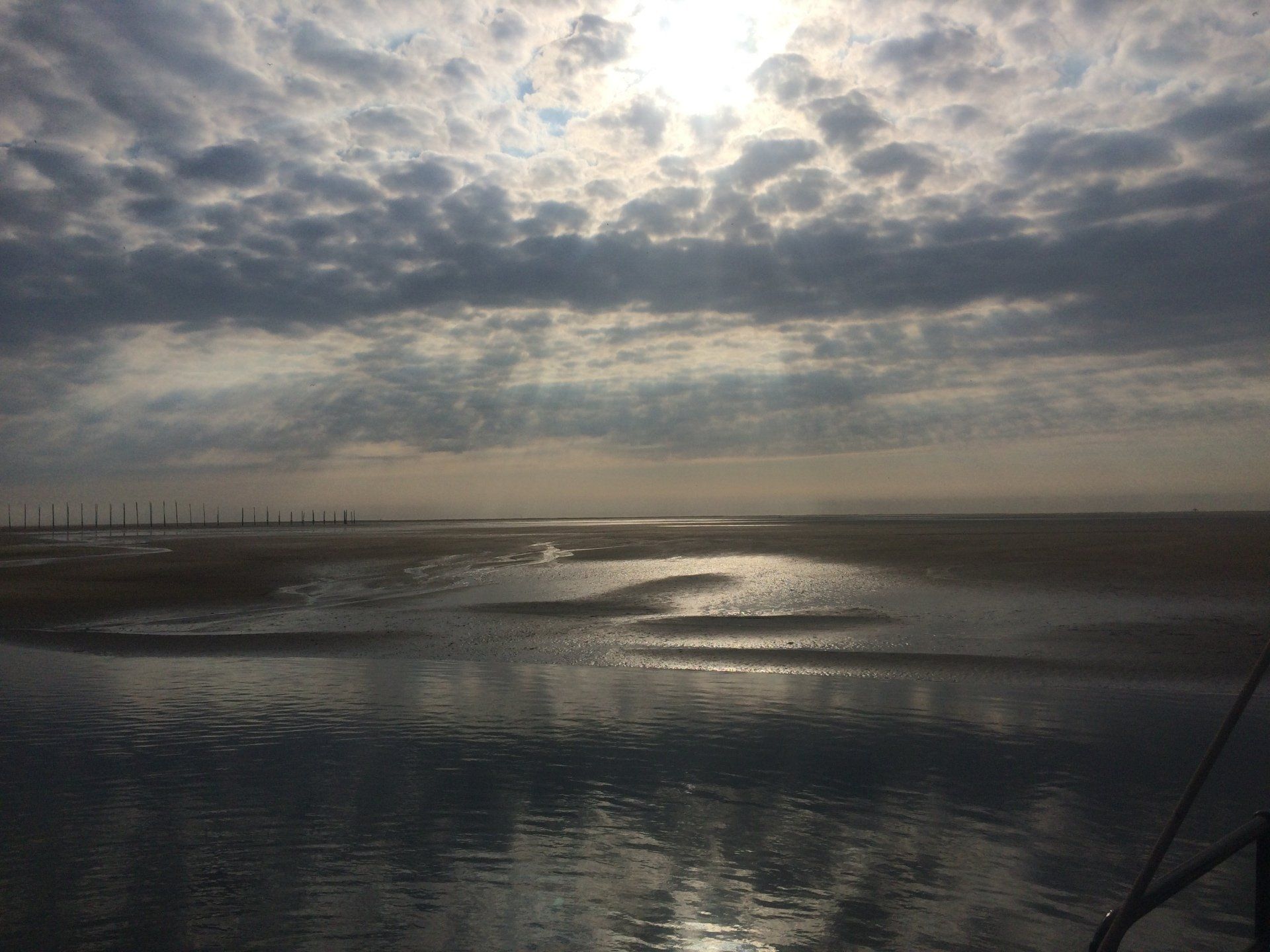 Bewölkter Himmel über einer ruhigen, nassen Sandbank; Sonne lugt durch die Wolken; Wasser im Vordergrund.
