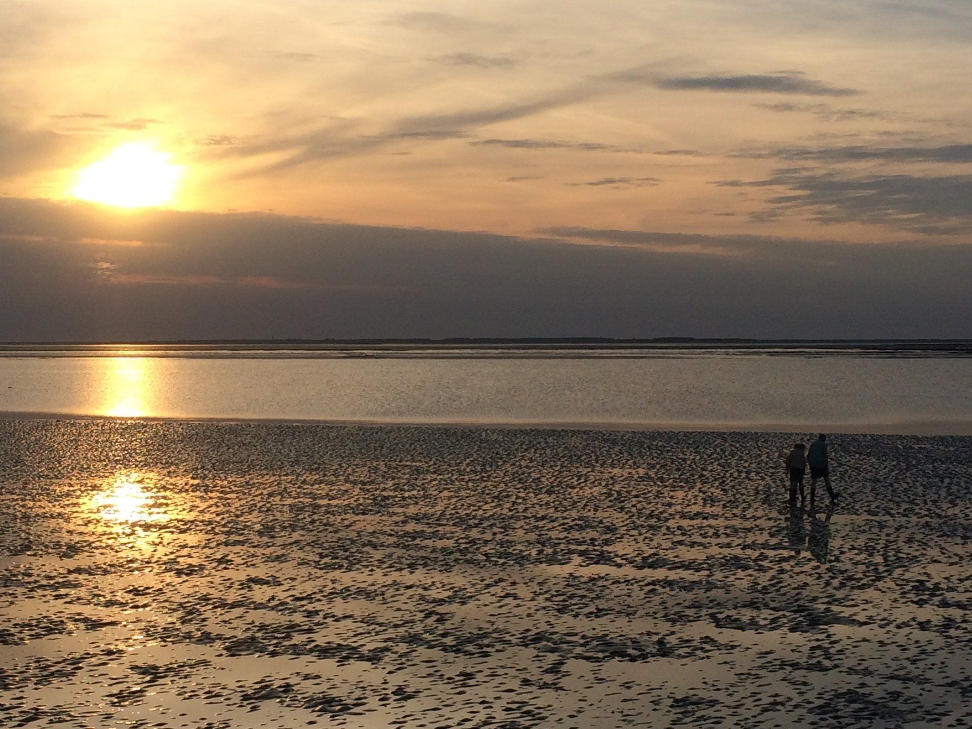 Sonnenuntergang über einem Strand, mit zwei in der Ferne spazierenden Gestalten, die Sonne spiegelt sich im nassen Sand.
