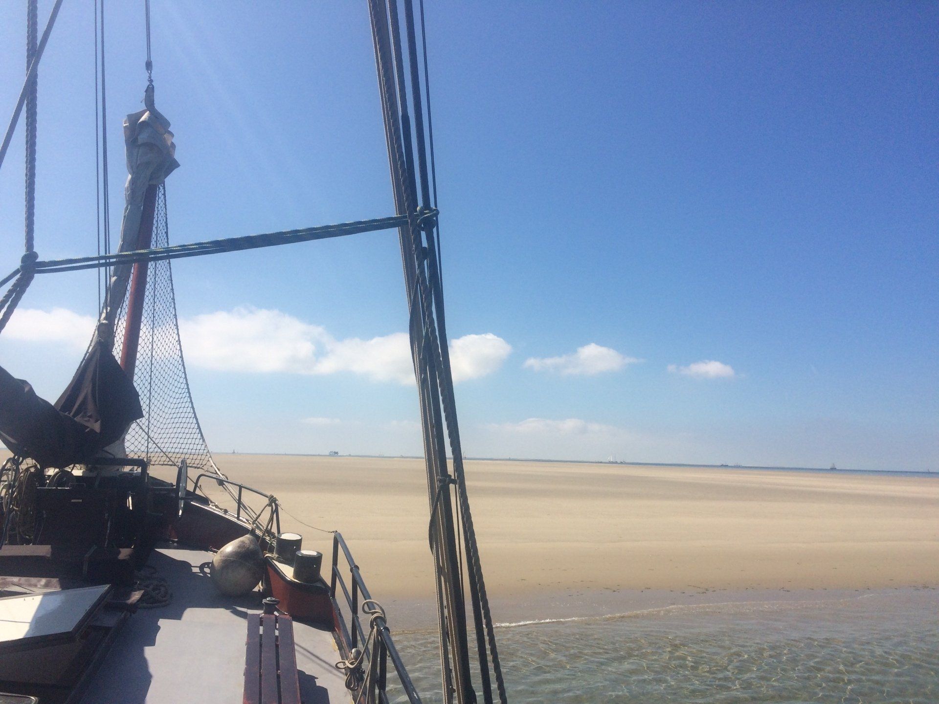 Blick vom Schiffsdeck auf einen weitläufigen Sandstrand unter strahlend blauem Himmel mit vereinzelten Wolken.