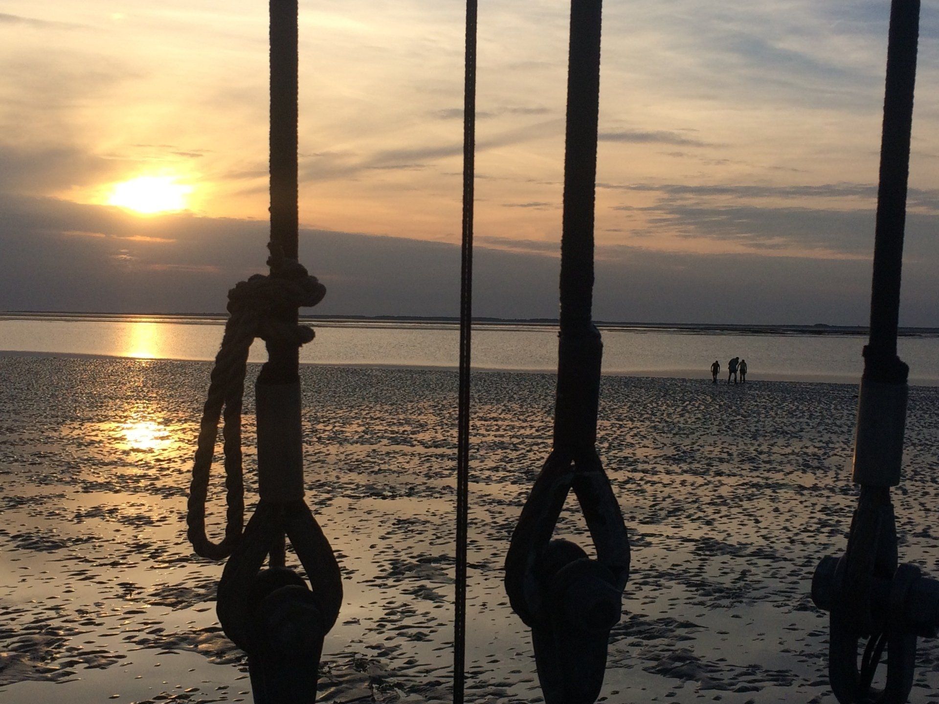 Sonnenuntergang über einem Strand, durch Takelageseile betrachtet. Menschen laufen auf dem nassen Sand, die Sonne spiegelt sich darin.