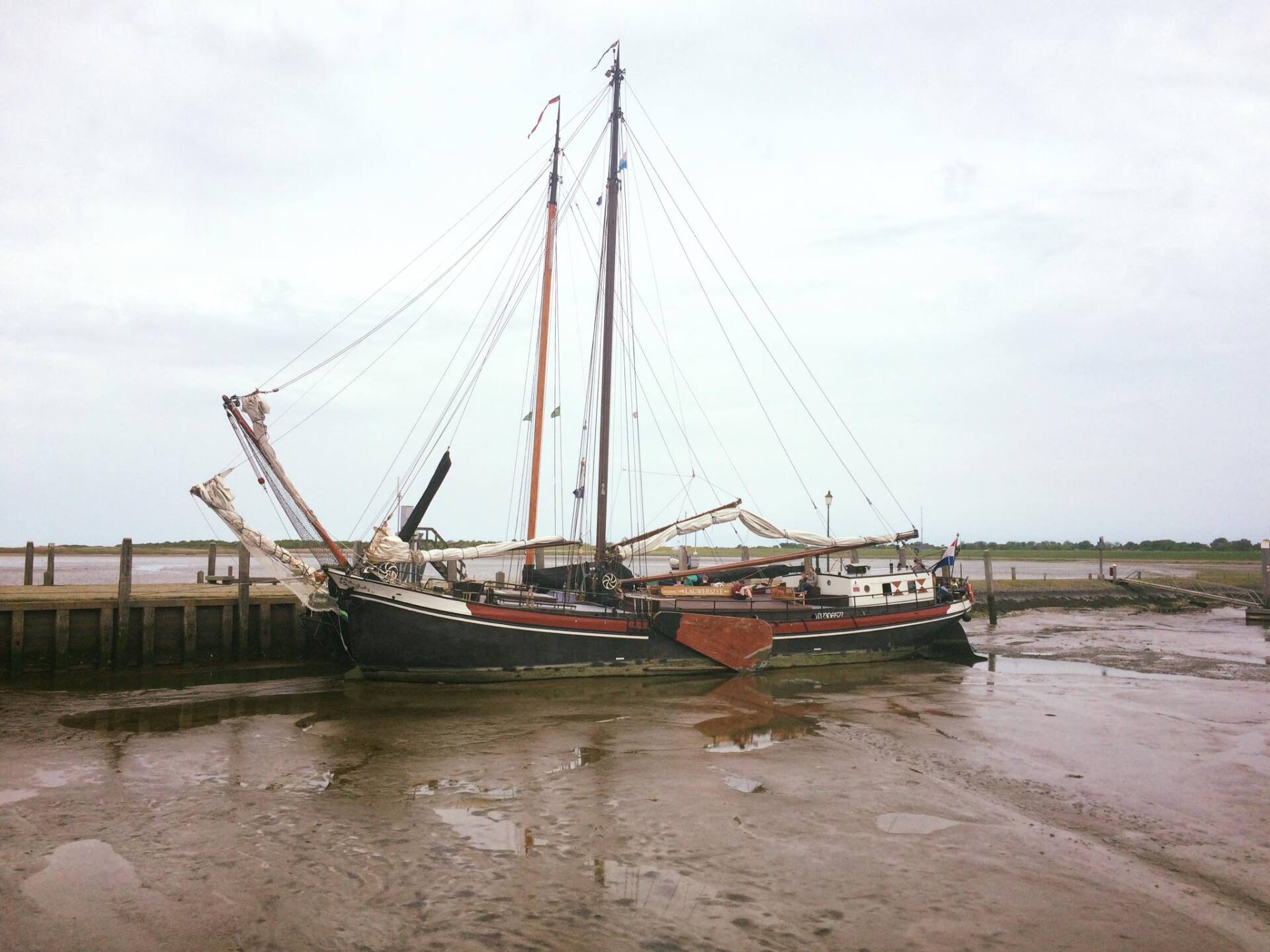 Ein Segelboot mit hohem Mast liegt unter einem bewölkten Himmel im Schlamm in der Nähe eines Piers.