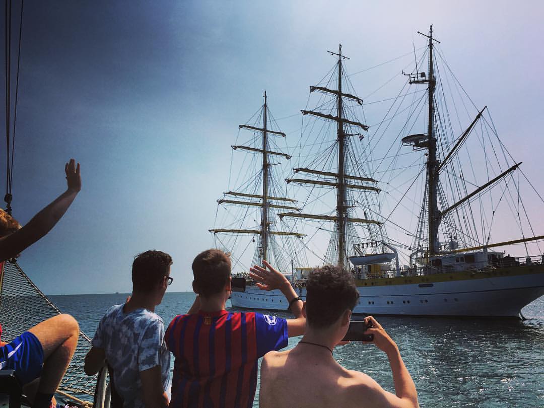 An einem sonnigen Tag winken Menschen auf einem Boot einem großen Segelboot zu.