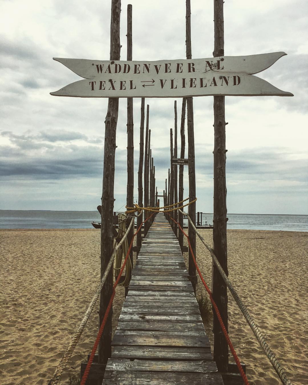 Holzstegschild mit Hinweis auf Texel und Vlieland an einem Sandstrand, bewölkter Himmel.