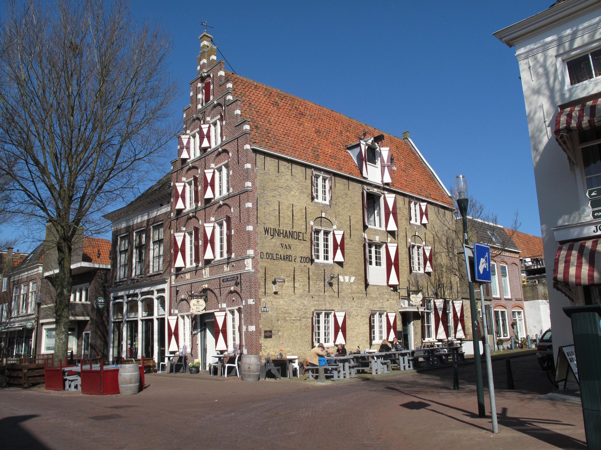 Historisches Backsteingebäude in Harlingen mit roten Fensterläden, Restauranttische draußen, blauer Himmel.