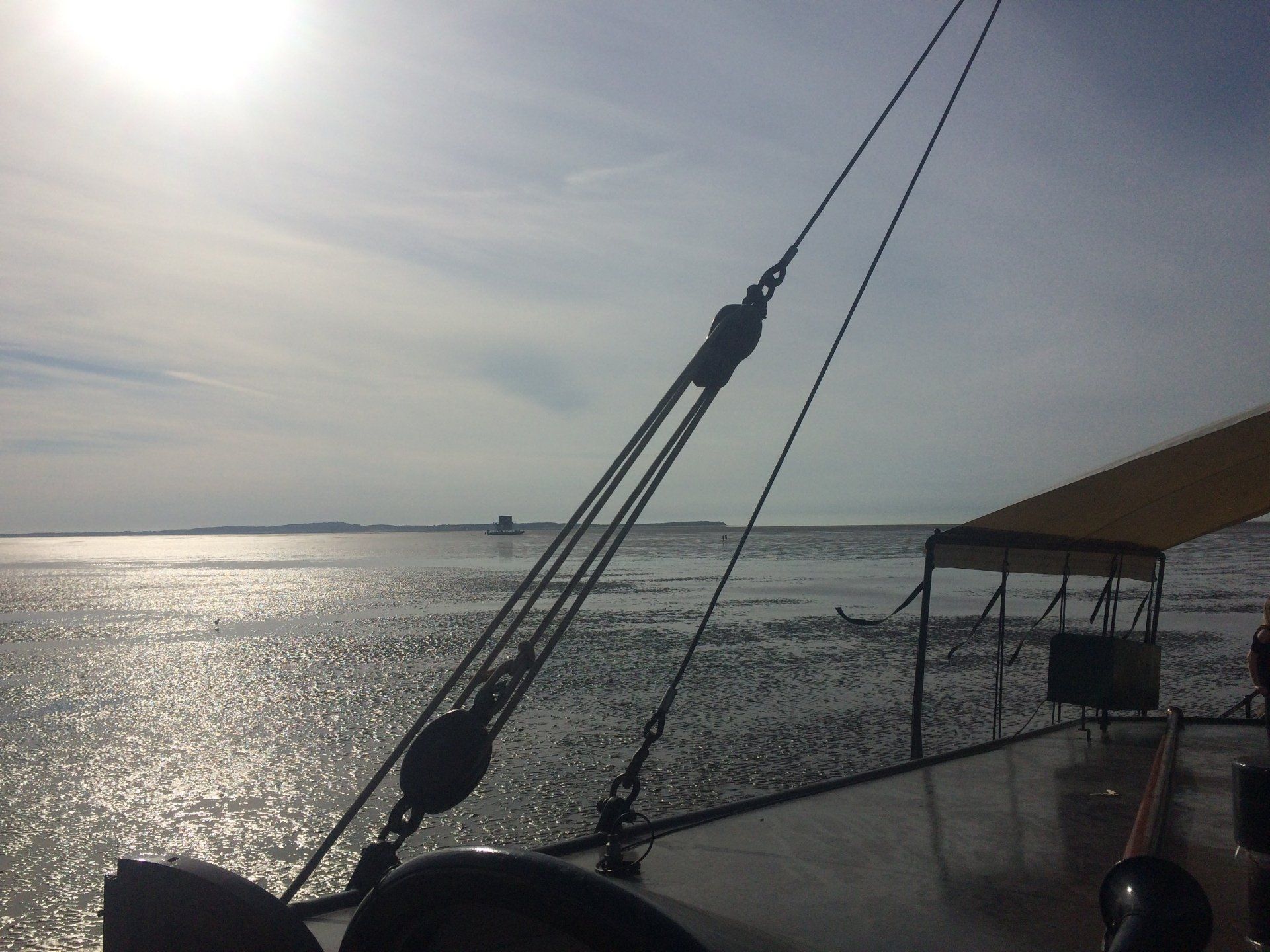 Sonnenlicht spiegelt sich auf dem Wasser. Die Takelage eines Schiffes zeichnet sich gegen den Himmel ab. In der Ferne ist eine kleine Insel zu sehen.