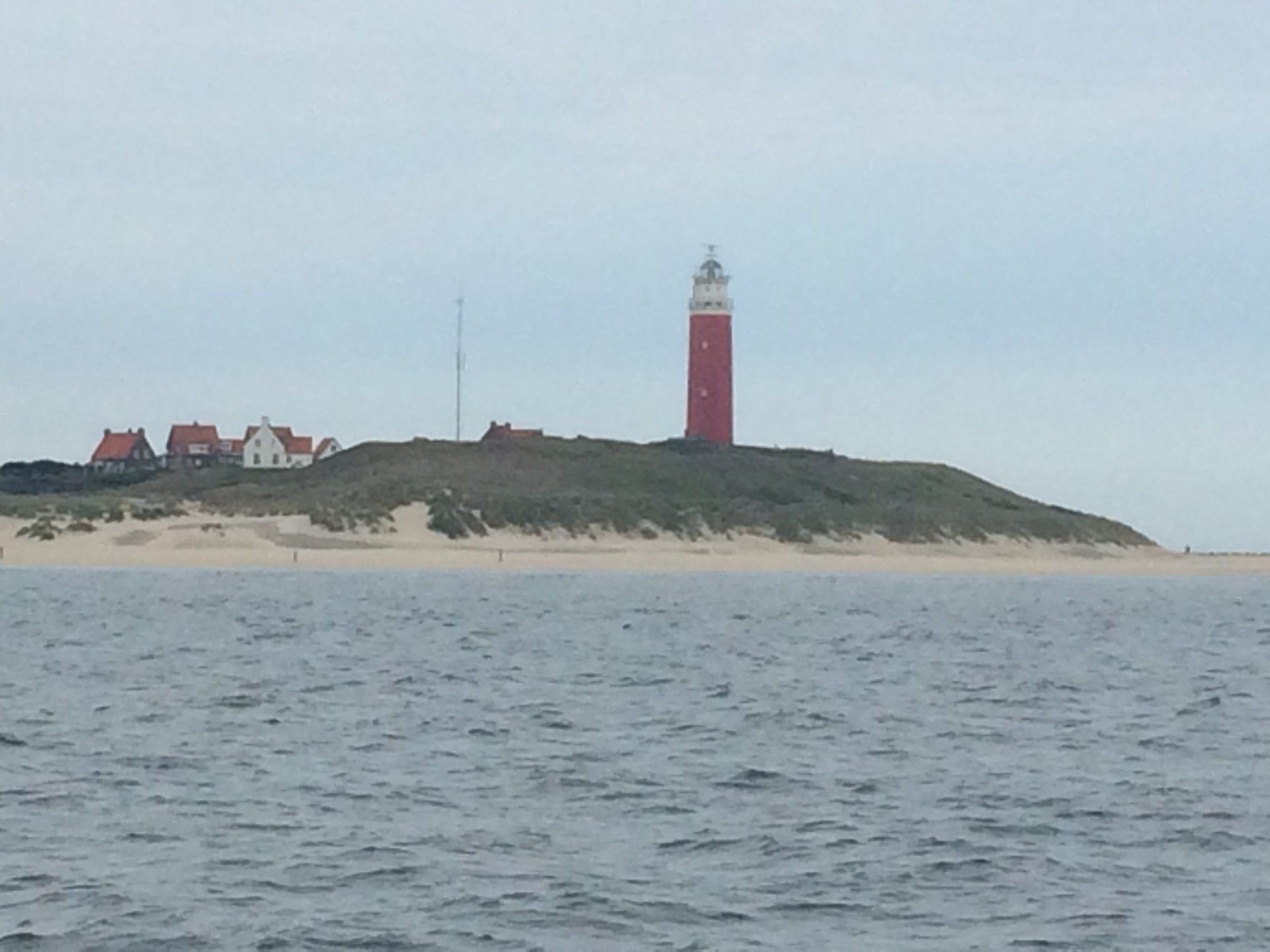 Rot-weißer Leuchtturm auf einer Düne mit Gebäuden, vom Meer aus unter bewölktem Himmel gesehen.