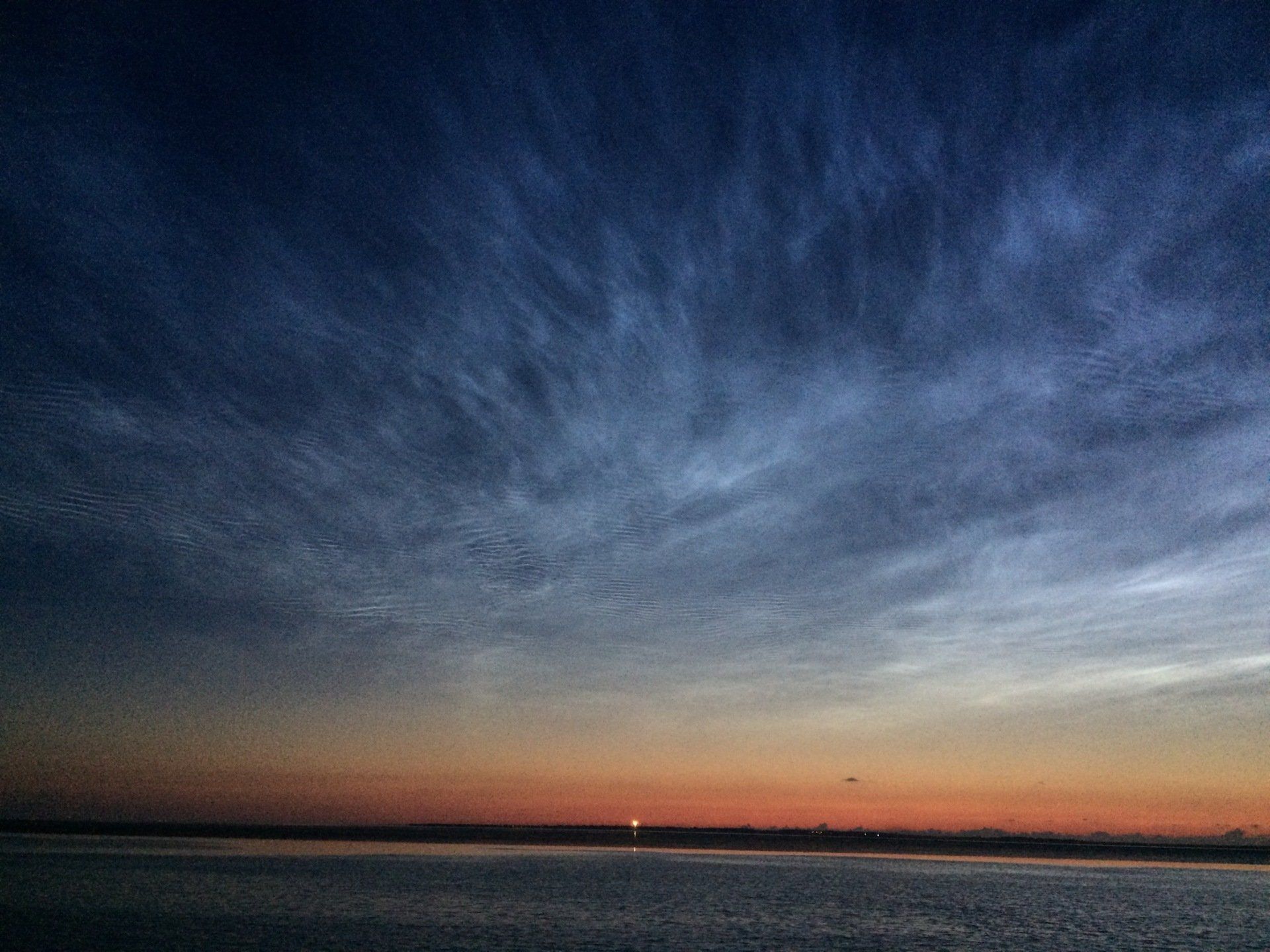 Leuchtende Nachtwolken, beleuchtet bei Sonnenuntergang, über einem ruhigen Horizont. Dunkelblaue und orange Farbtöne.