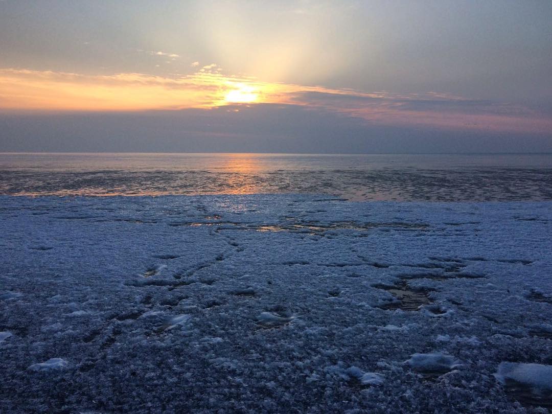 Sonnenaufgang über einem Gewässer mit Eis im Vordergrund, in Orange-, Blau- und Grautönen.