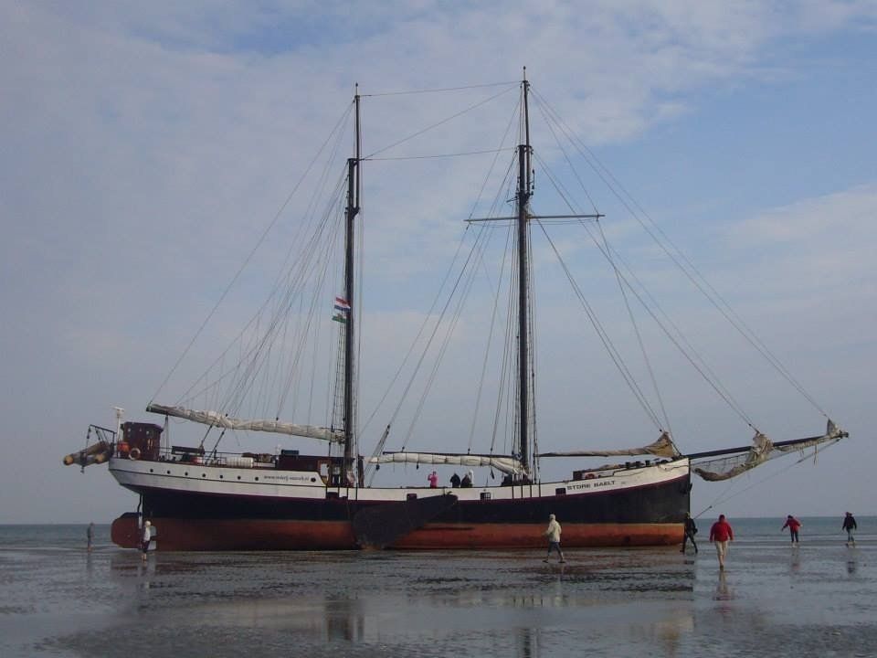 Großes Segelboot, das in seichtem Wasser auf Grund gelaufen ist, mit Menschen, die an einem bewölkten Tag in der Nähe spazieren gehen.