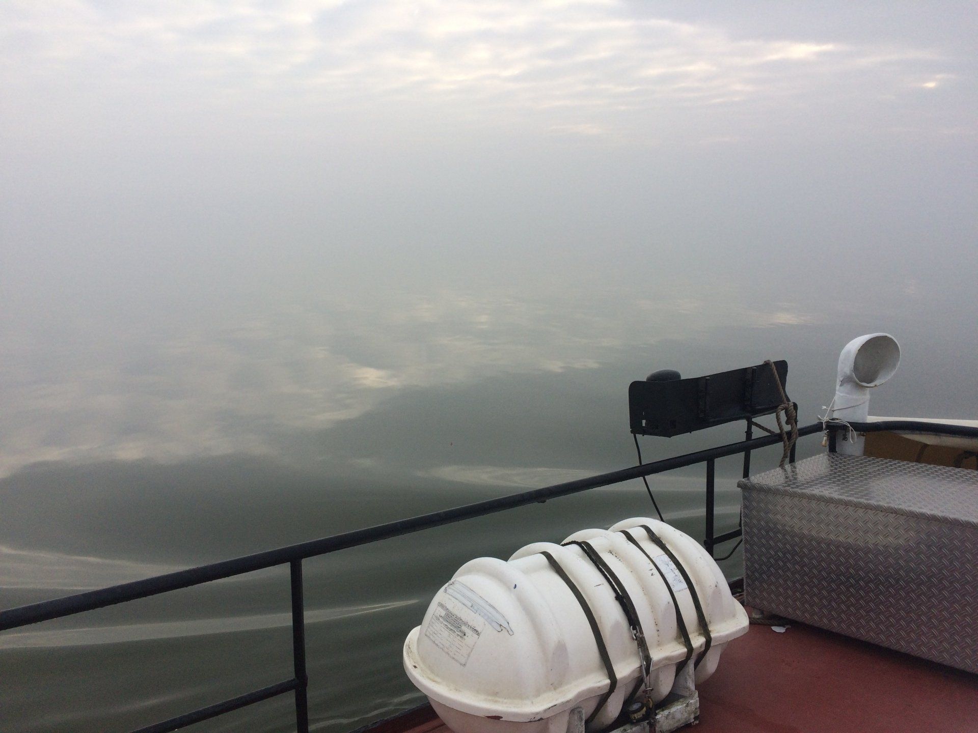 Blick vom Bootsdeck auf Wasser und nebligen Himmel. Auf dem Deck sind ein Rettungsfloß und Ausrüstung zu sehen.