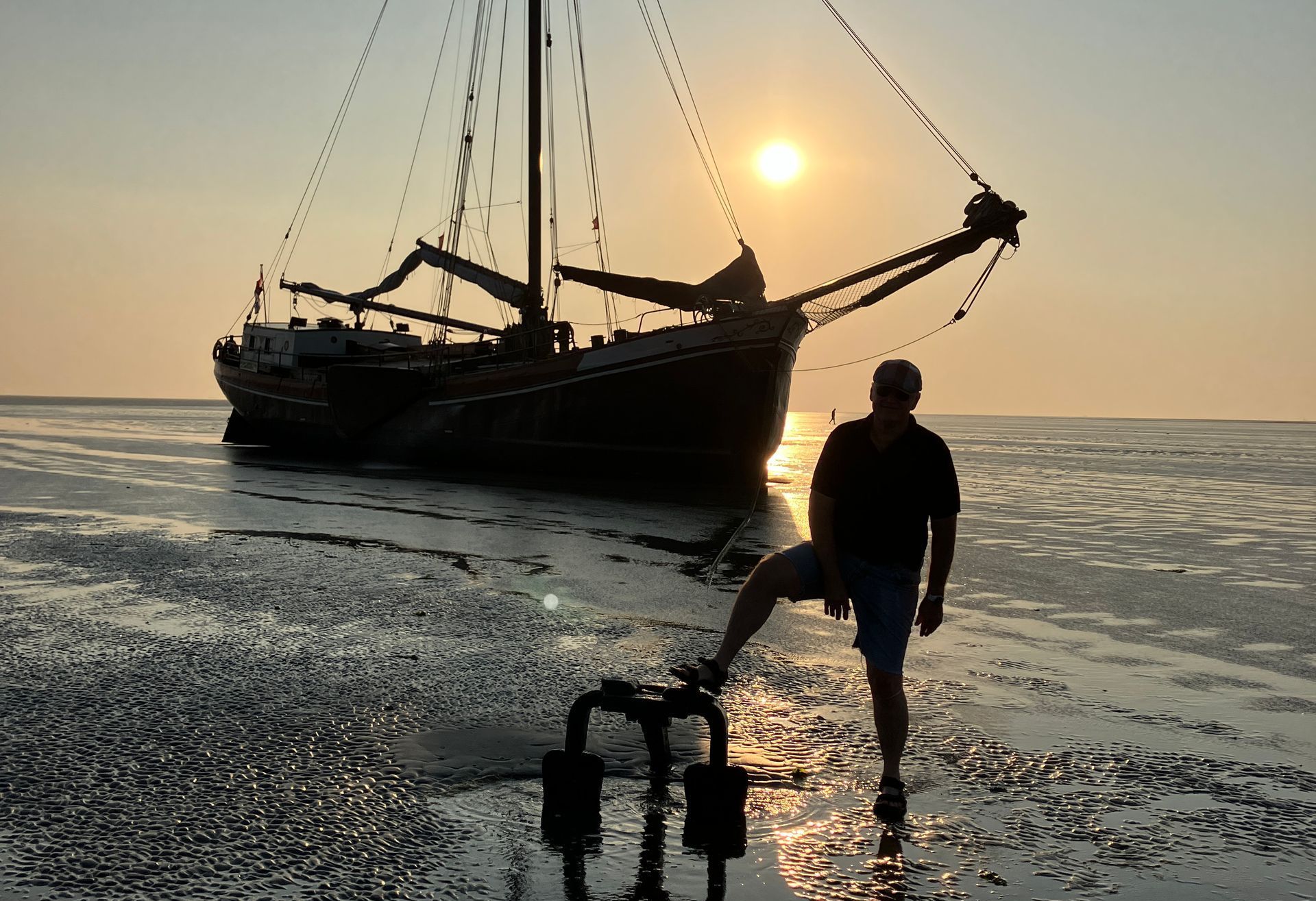 Mann posiert mit einem trockengefallenen plattbodenschiff, das bei Sonnenuntergang auf dem Sandbank liegt und sich als Silhouette auf nassem Sand abzeichnet.
