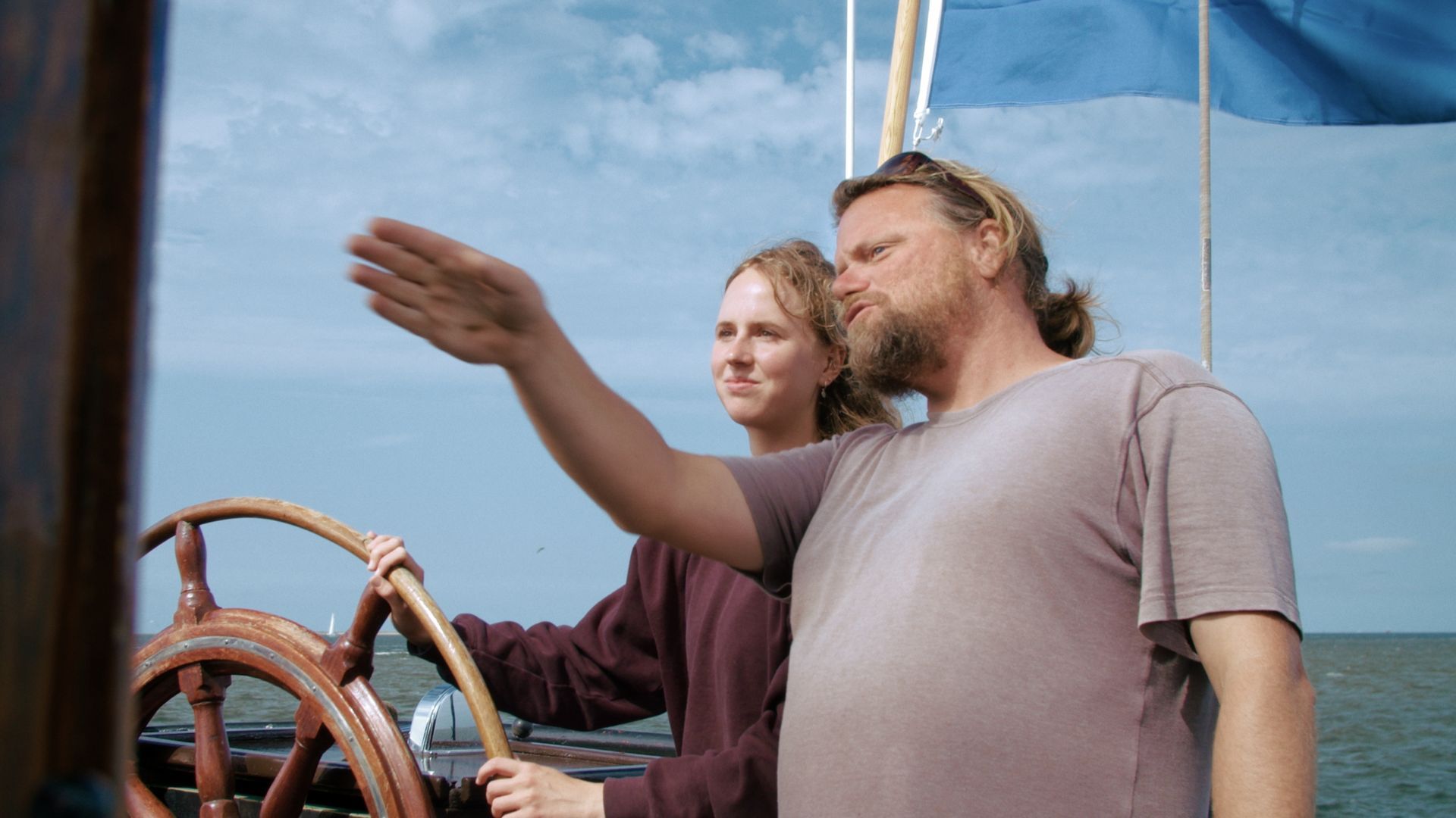 Man wijst naar de horizon terwijl hij een zeilboot bestuurt; vrouw staat naast hem.