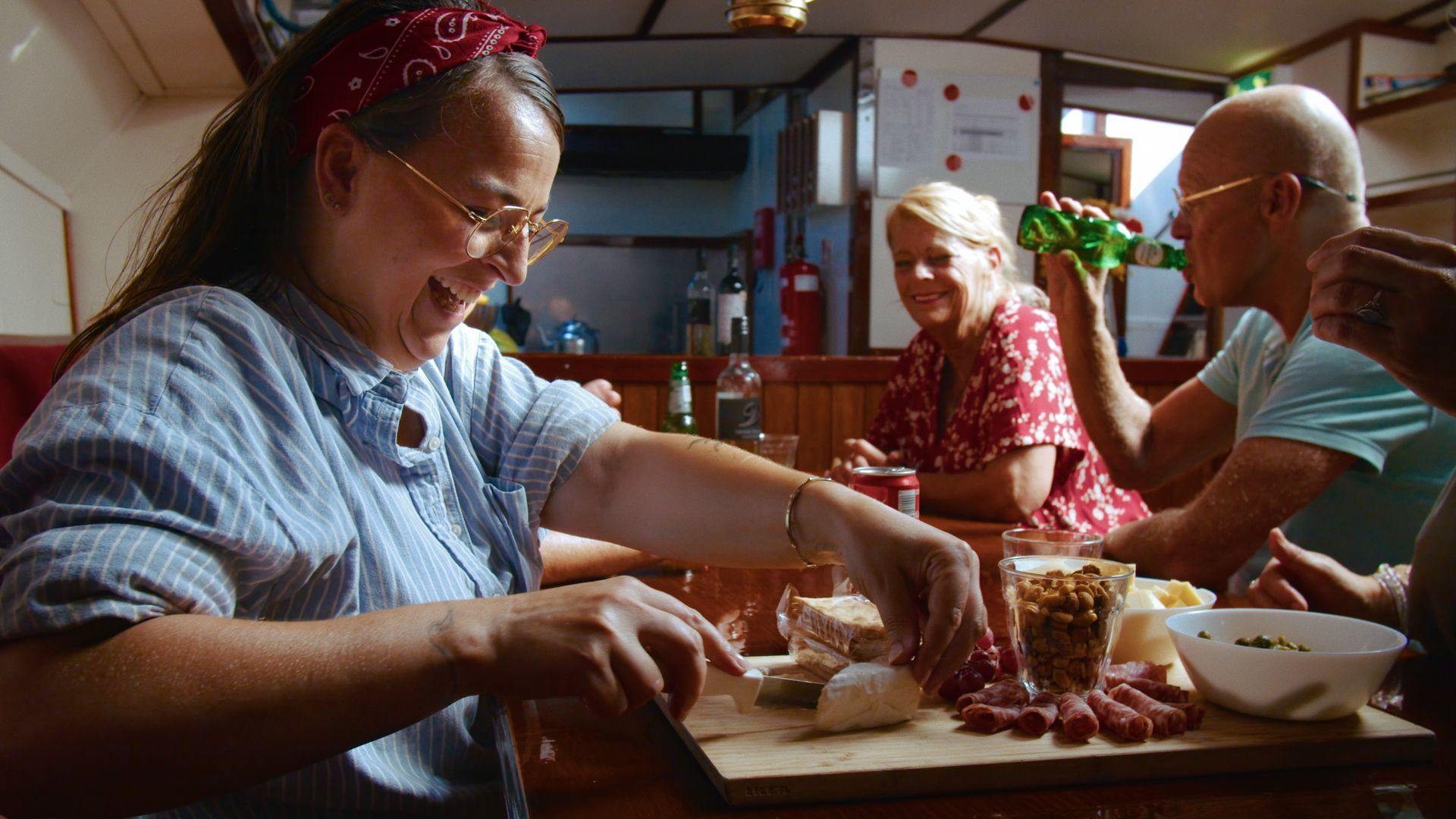 Vrouw snijdt eten op een boot met vrienden, lachend. Houten snijplank, bril, rode bandana.