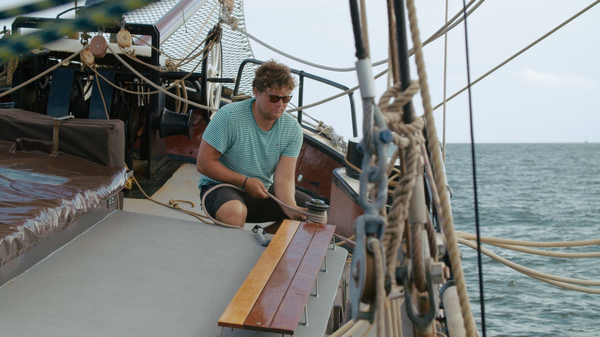 Man knielt op het dek van een boot en maakt een houten reling vast met een touw vlak bij het water.