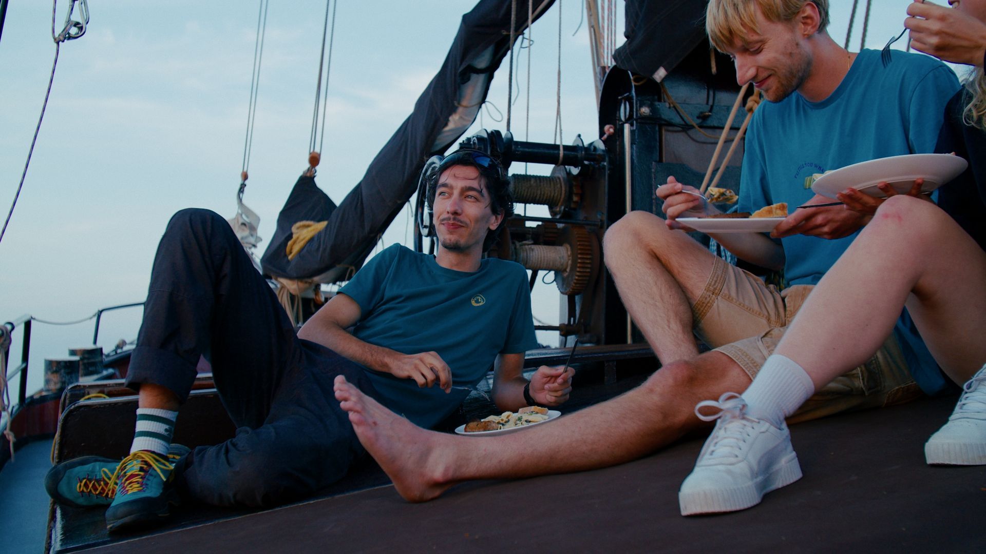 Mensen eten op een bootdek bij schemering. Eén ligt achterover, twee anderen eten noedels met stokjes.