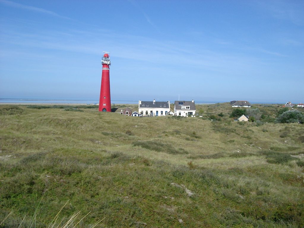 Roter Leuchtturm von Schiermonnikoog und weiße Gebäude auf einer grasbewachsenen Düne unter einem klaren blauen Himmel.