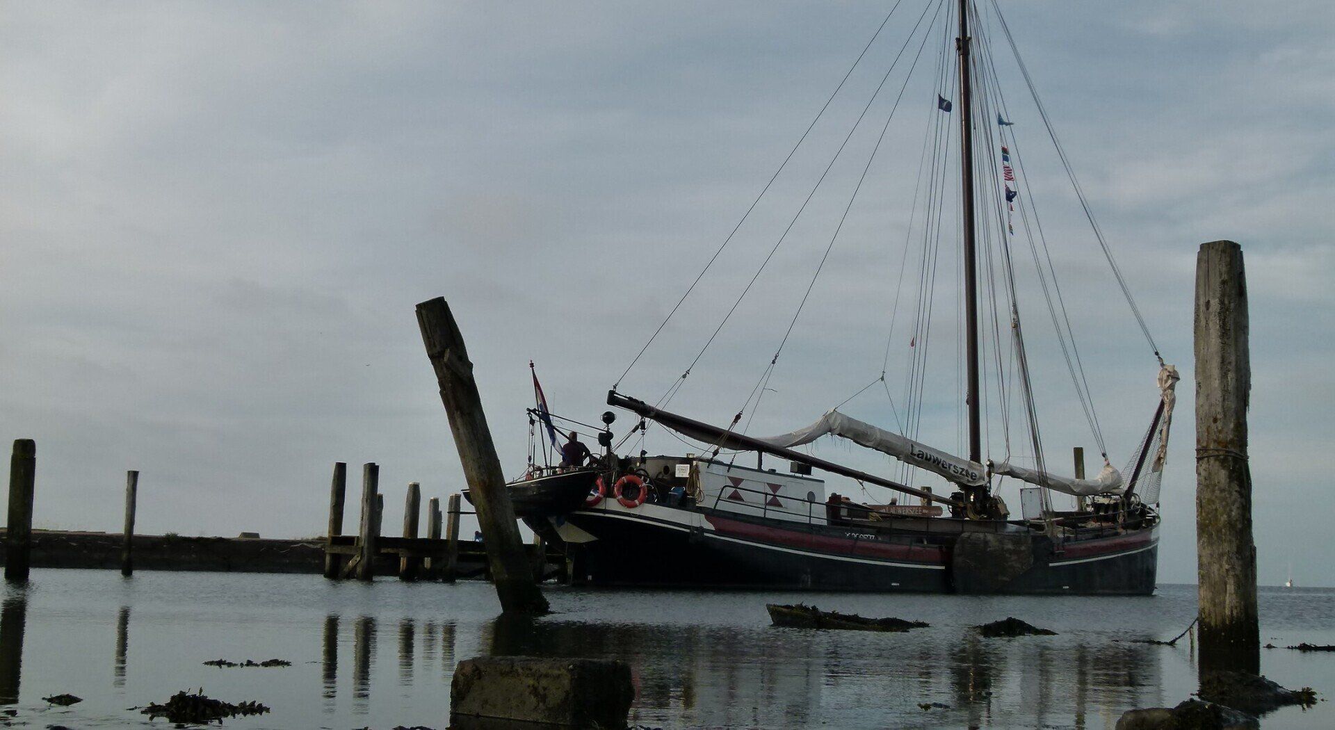Ein verwittertes Segelboot an einem Pier, brauner Rumpf, Masten mit Seilen, bewölkter Himmel, trübes Wasser.