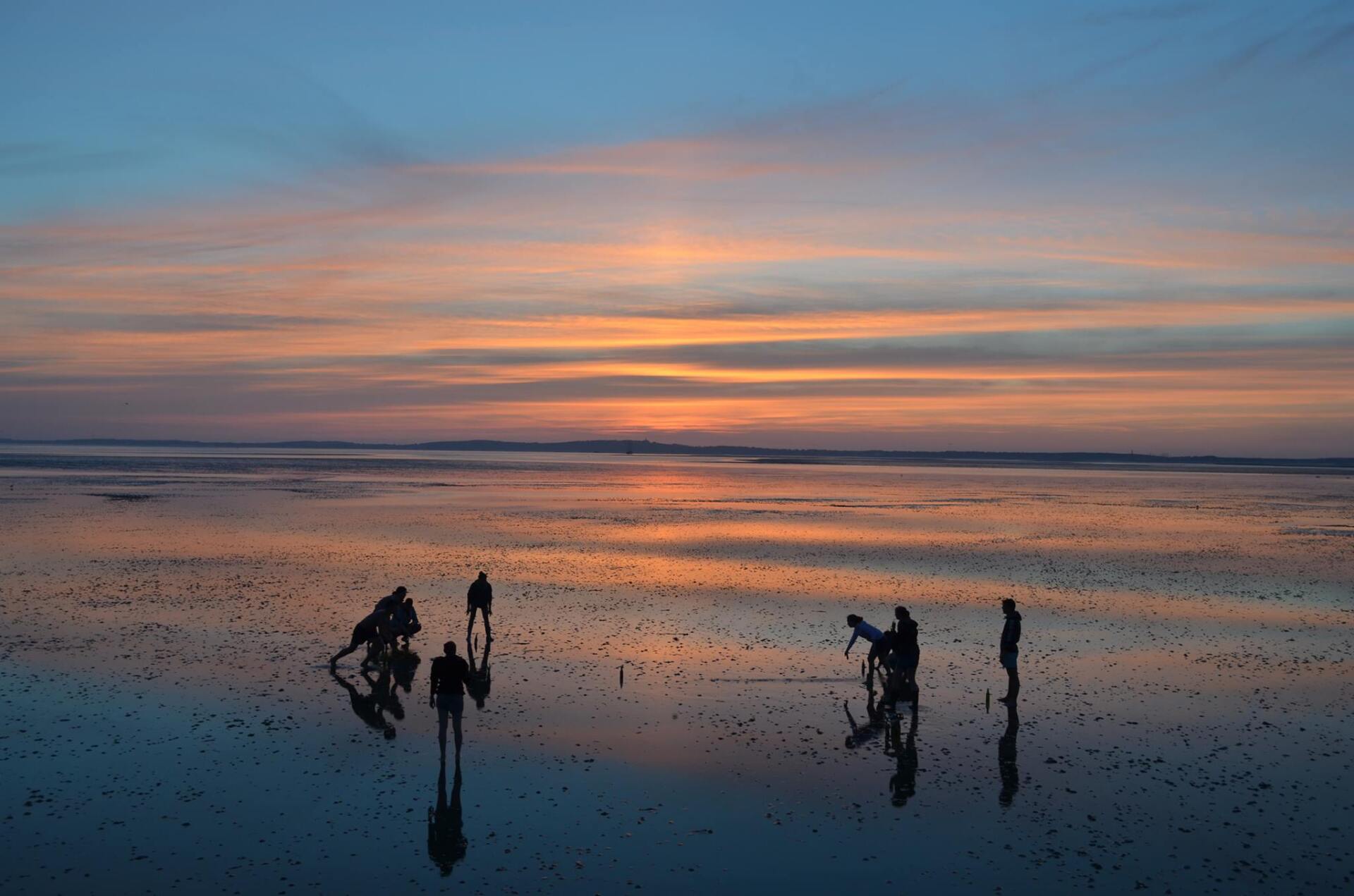 Sonnenuntergang über einer spiegelnden tockengefallen Sandbank auf dem Wattenmeer. Silhouetten von Figuren stehen am Wasserrand; der Himmel ist in orange, blauen und rosa Farbtönen gehalten.