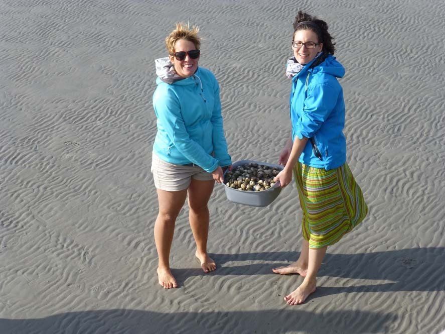 Zwei Frauen am Strand, die einen Korb mit Muscheln halten. Sie tragen blaue Jacken und sind barfuß.