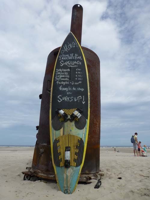 Ein Surfbrett als Speisekarte an Strand von Schiermoinnikoog, vor einer rostigen Metallkonstruktion platziert; ein sonniger Tag.