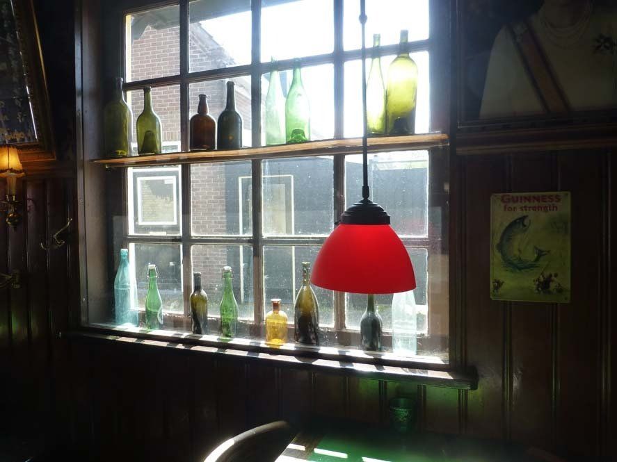 Ein Fenster mit Regalen, auf denen verschiedene Glasflaschen, eine rote Lampe und ein Guinness-Poster stehen. Kneipe Zwaantje auf West-Terschelling
