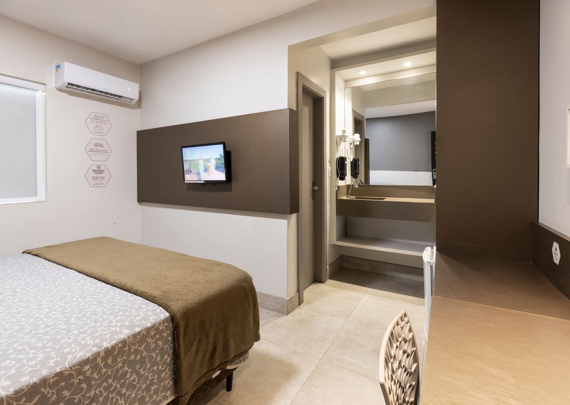 Quarto de hotel compacto com cama, TV de parede, ar condicionado e lavatório no banheiro.