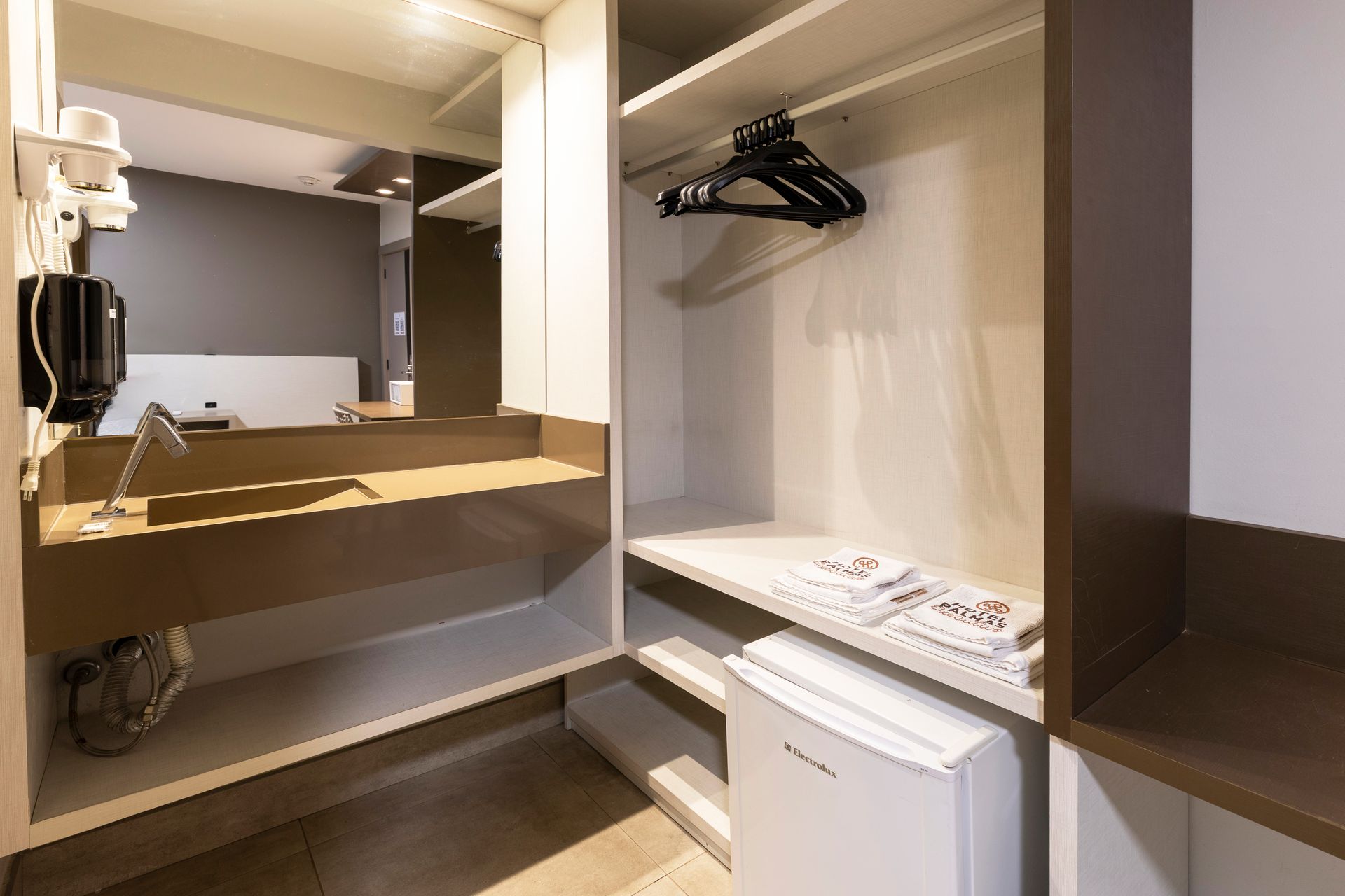 Quarto de hotel compacto com escrivaninha, espelho, cabides e armário aberto.