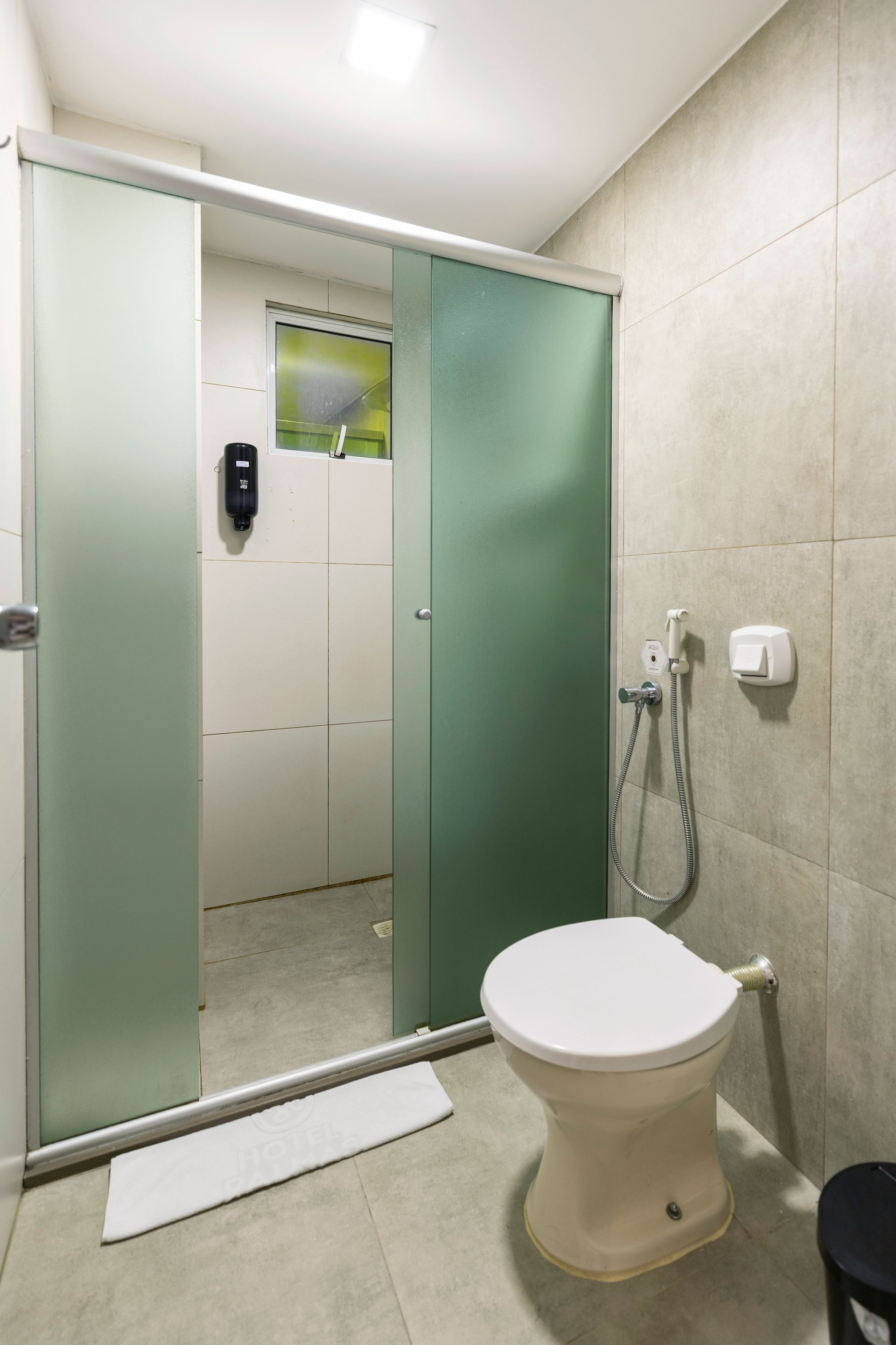 Banheiro pequeno com box de vidro fosco, ducha higiênica fixada na parede e paredes revestidas com azulejos bege.