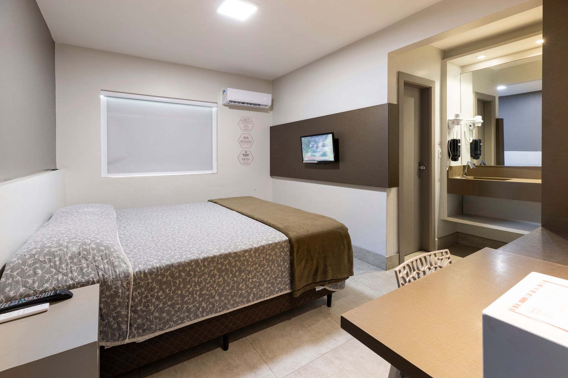 Quarto de hotel moderno com cama, TV de parede, escrivaninha e persianas.