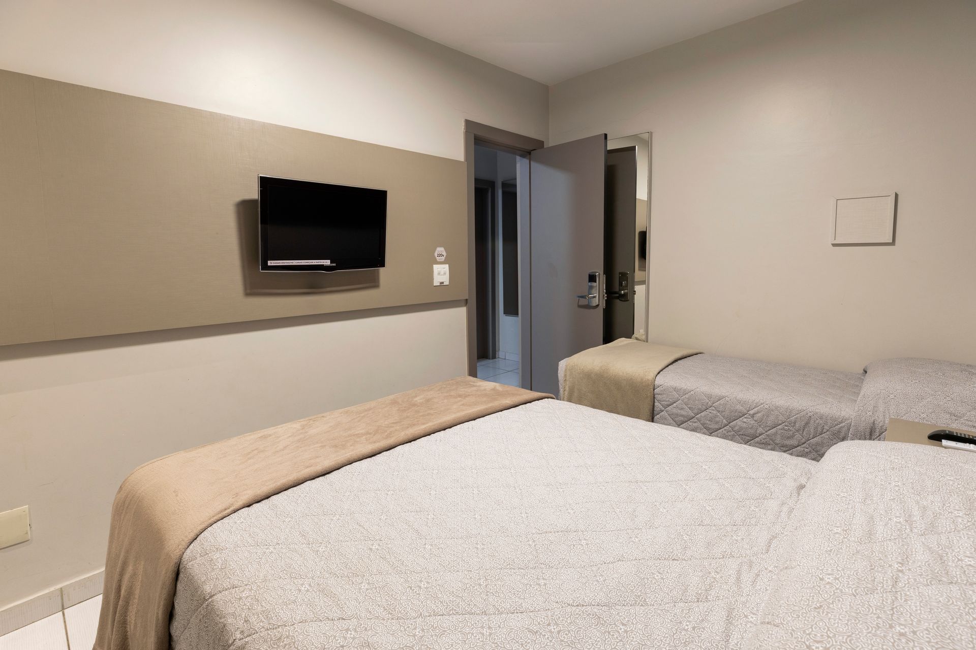Quarto de hospital com duas camas arrumadas, TV de parede e porta aberta para um corredor.