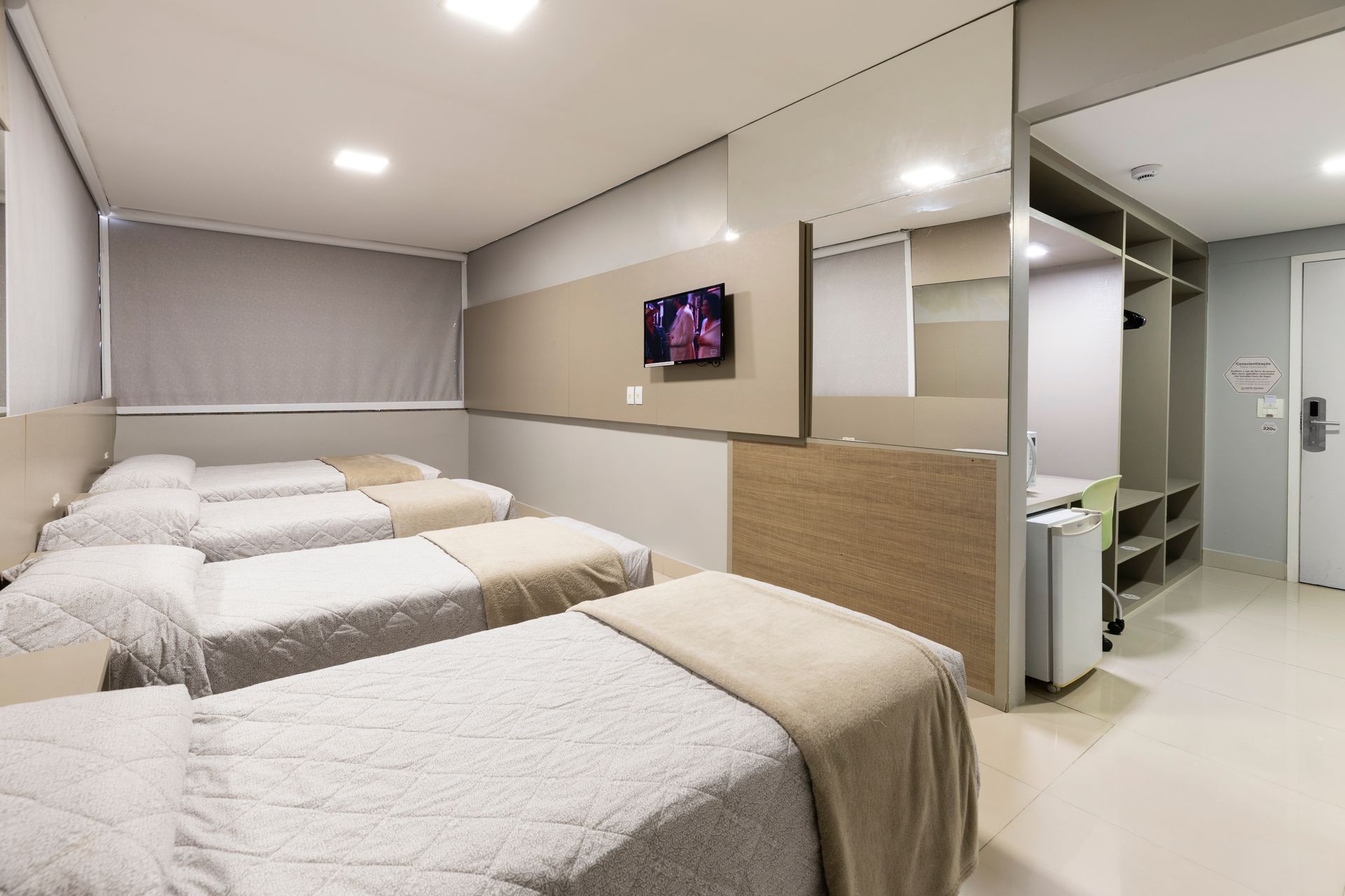 Quarto de dormitório com quatro camas de solteiro arrumadas, paredes bege e uma TV fixada na parede.