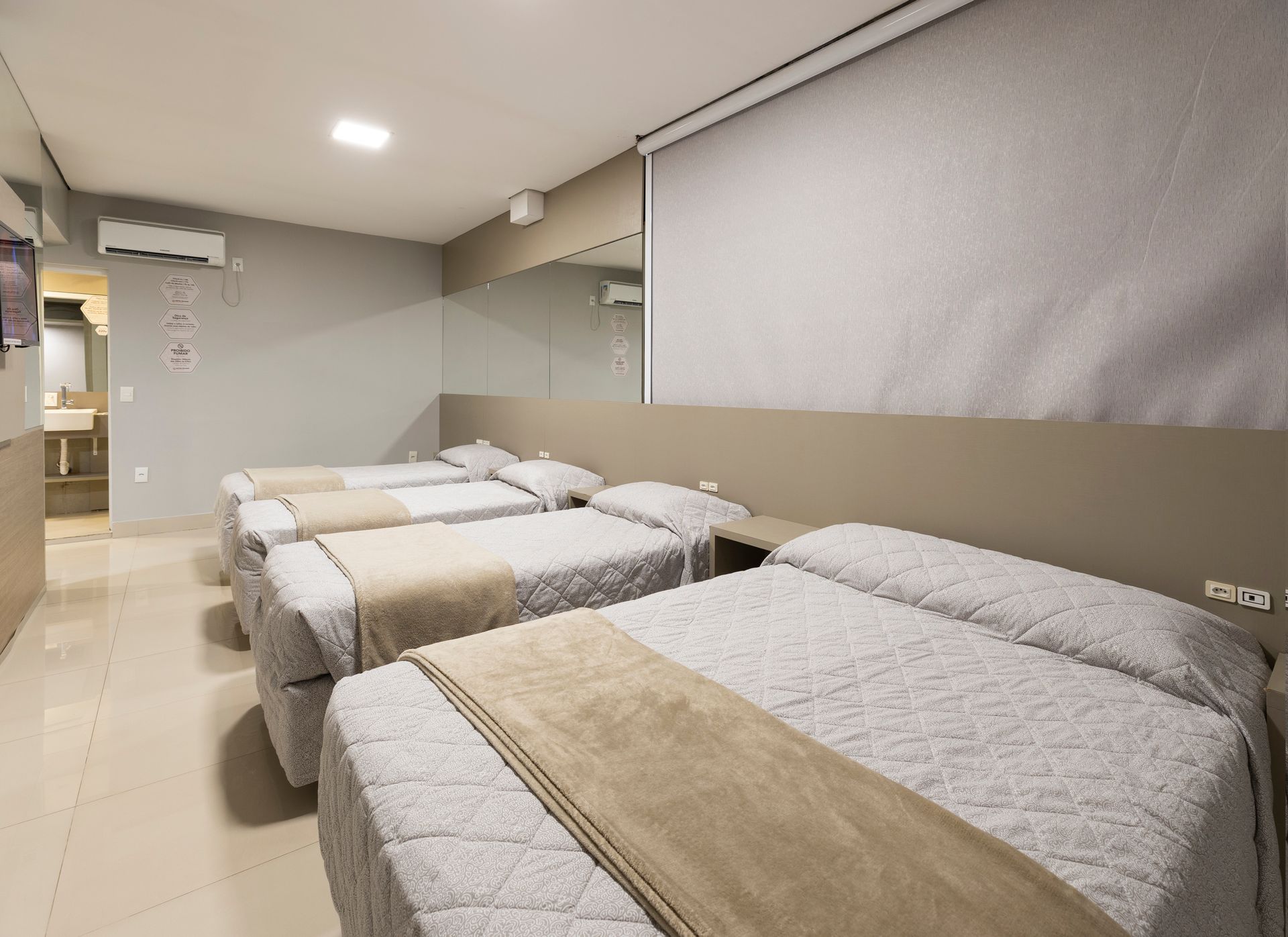 Quarto de hotel espartano com três camas de solteiro arrumadas e cortinas cinzentas contra uma parede bege.