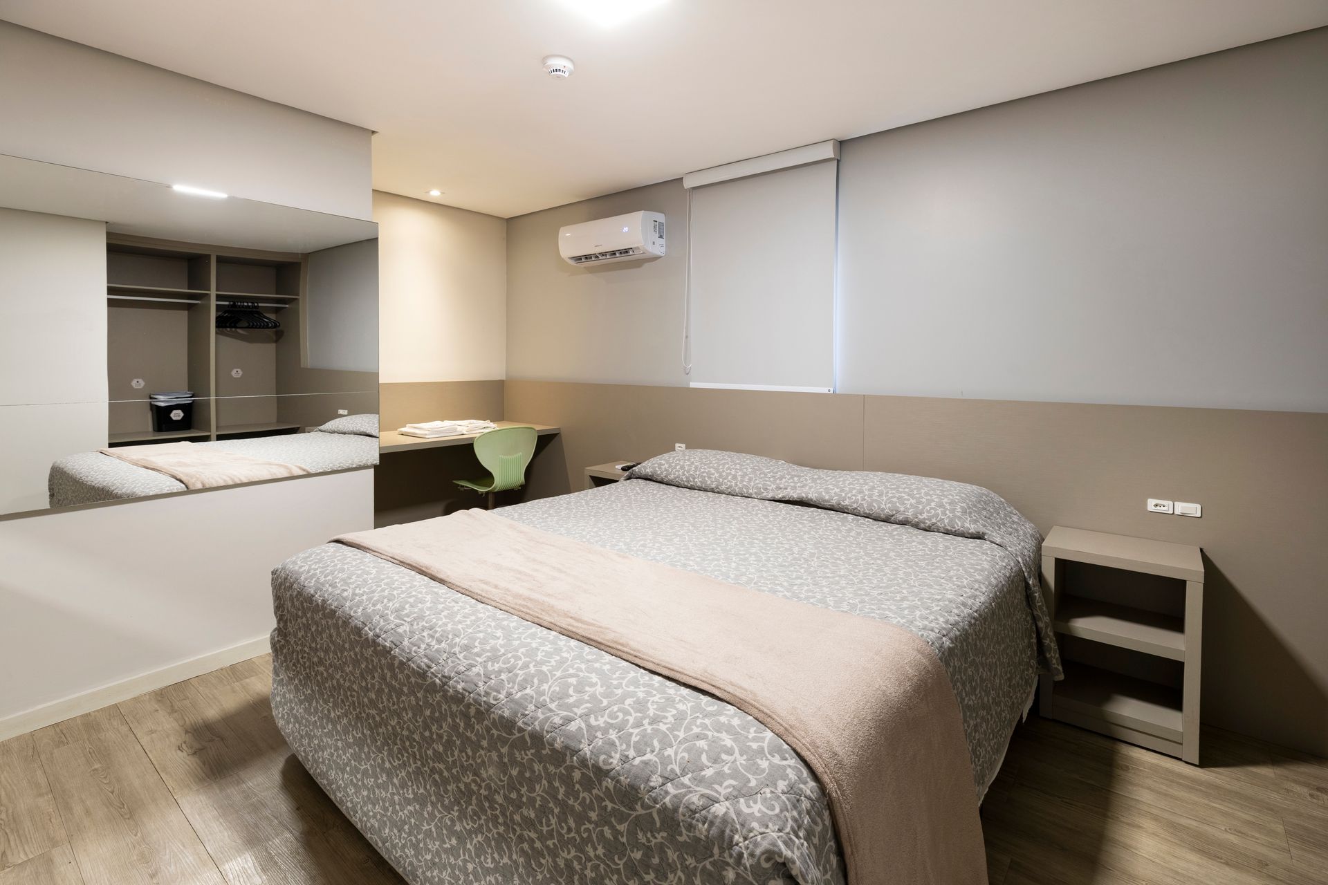 Quarto moderno com duas camas, decoração neutra, grande espelho de parede e ar condicionado instalado acima da janela.