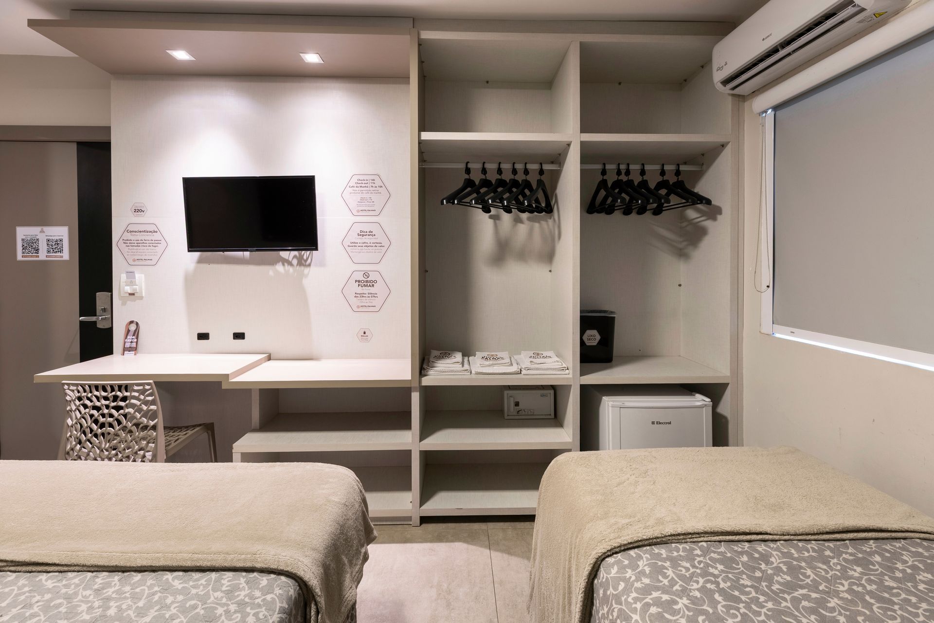 Quarto de hotel com duas camas, TV de parede, escrivaninha, armário aberto e persianas.
