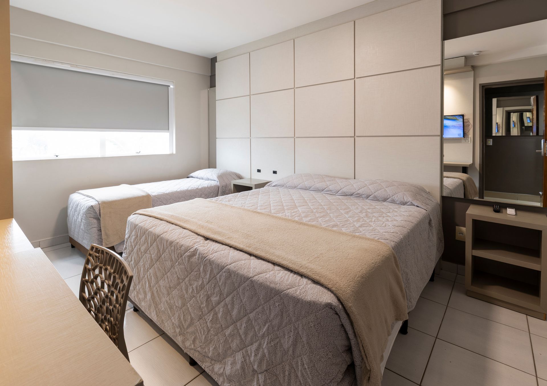 Quarto de hotel com duas camas bem arrumadas, uma escrivaninha e uma grande janela com persiana.